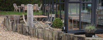 Un espacio al aire libre con mesas y sillas de madera, una planta pequeña en maceta y un recinto de vidrio con un cartel de clasificación de higiene alimentaria.