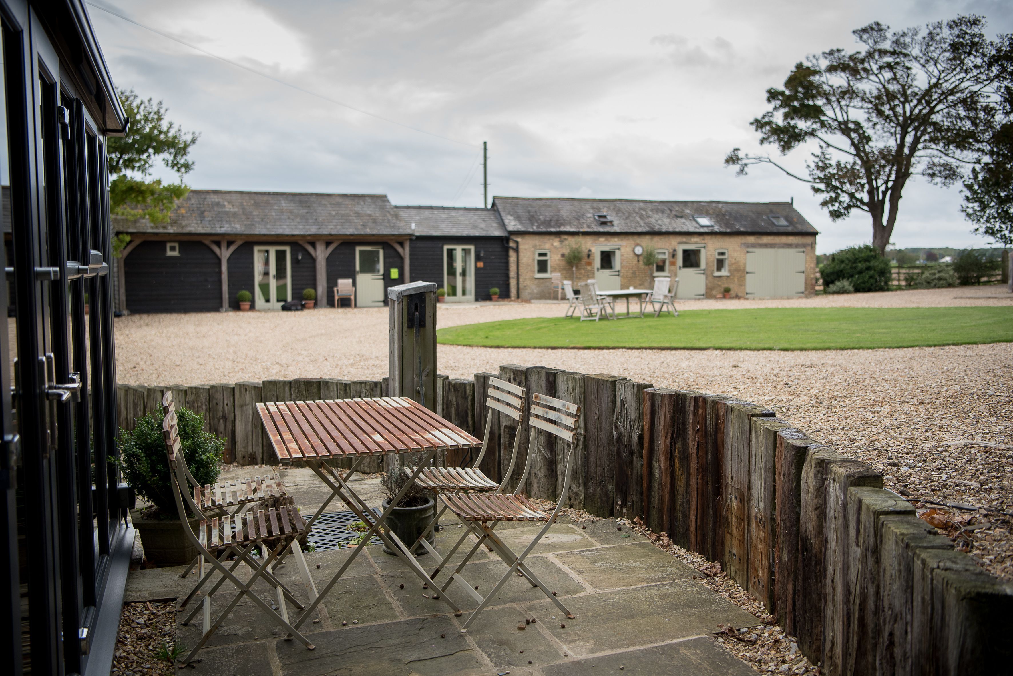 An outdoor patio with a wooden table and chairs, a gravel path, and a building with multiple doors and windows in the background.