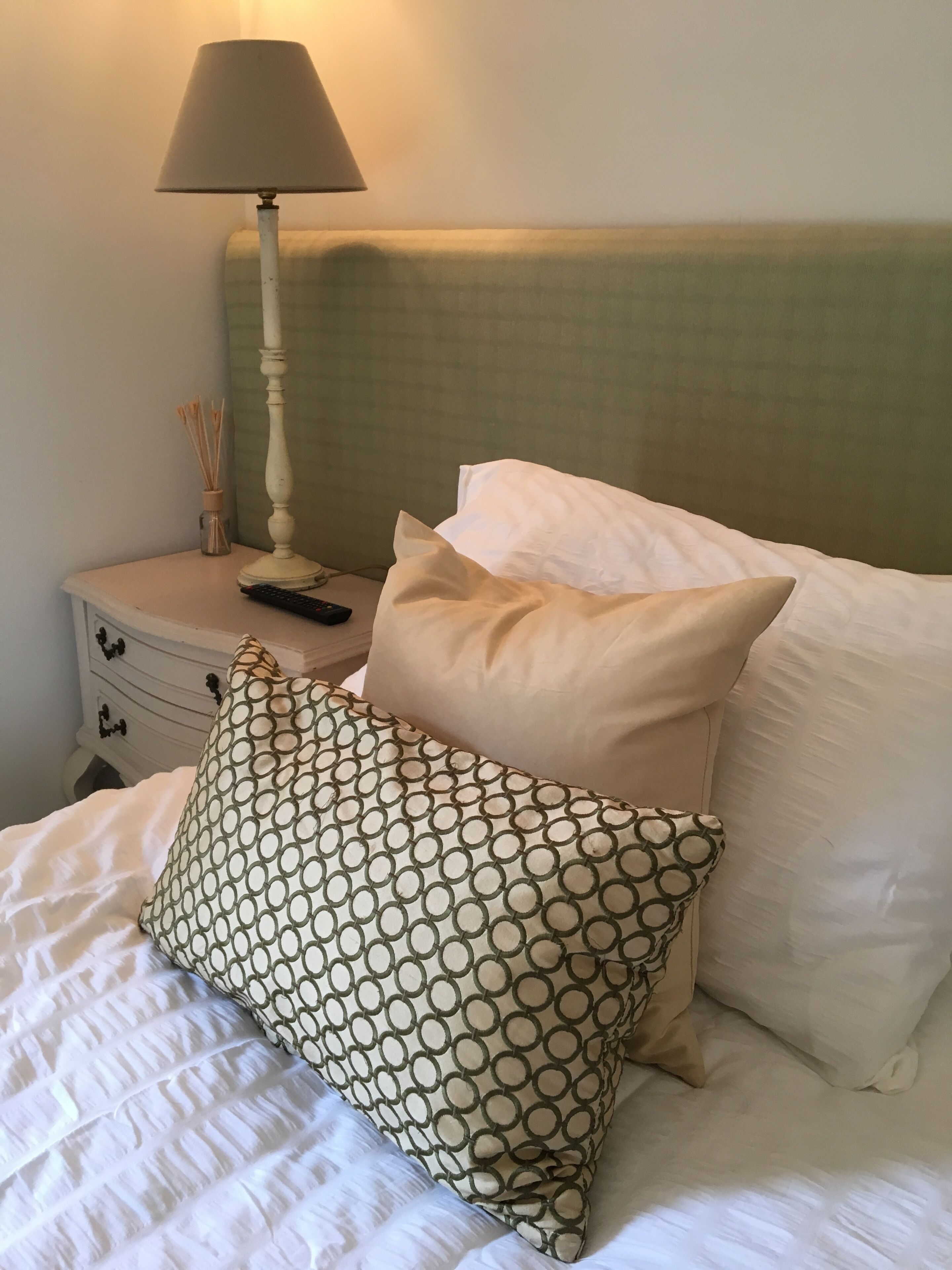 Un letto rifatto con cuscini fantasia e lisci, una lampada da comodino e un piccolo tavolino con un telecomando e un vaso di canne.
