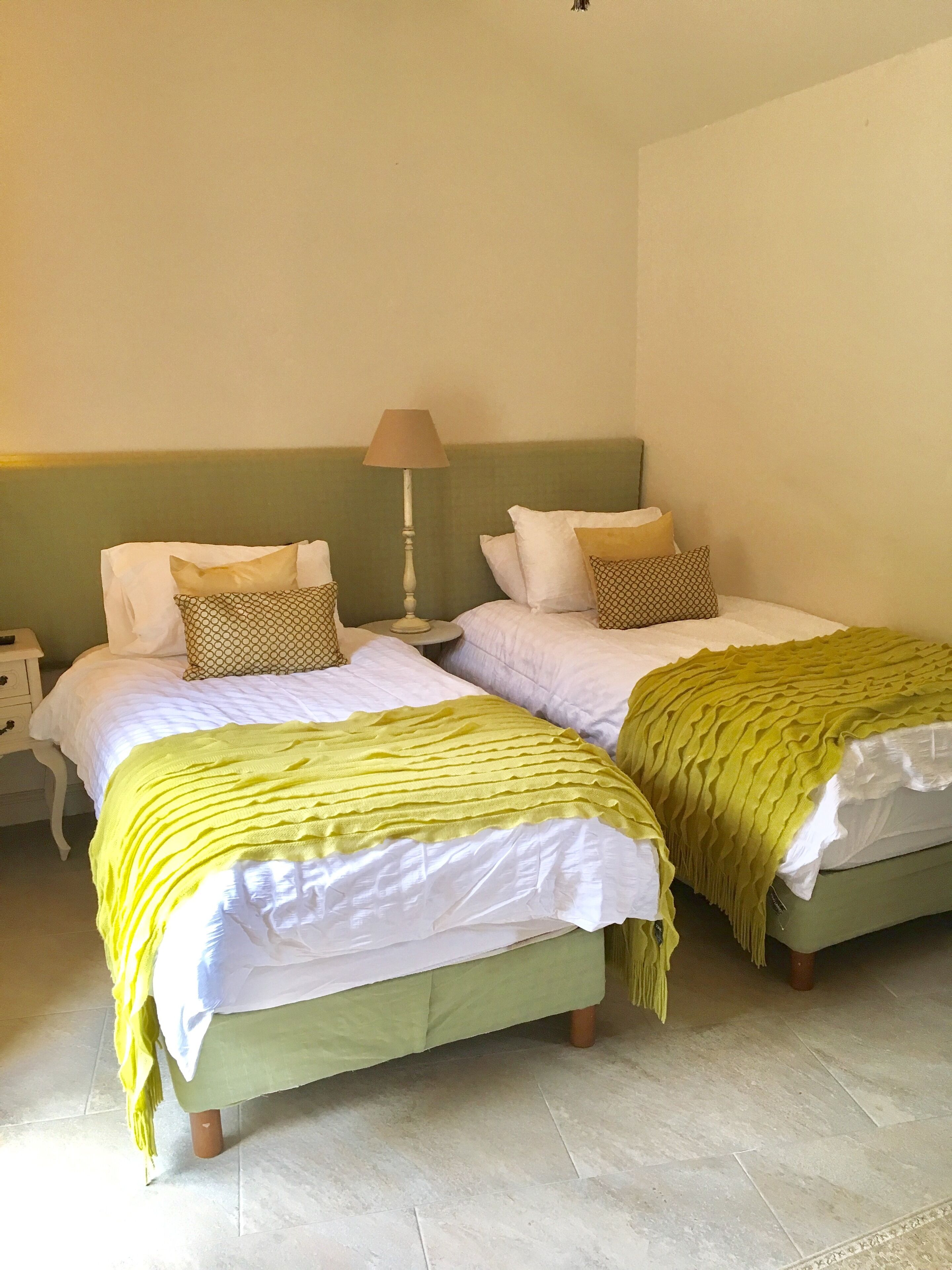 Ein Zimmer mit zwei Betten, jeweils mit einer grünen Tagesdecke und weißen Bettwäsche. An einer Seite steht ein Nachttisch mit einer Lampe.