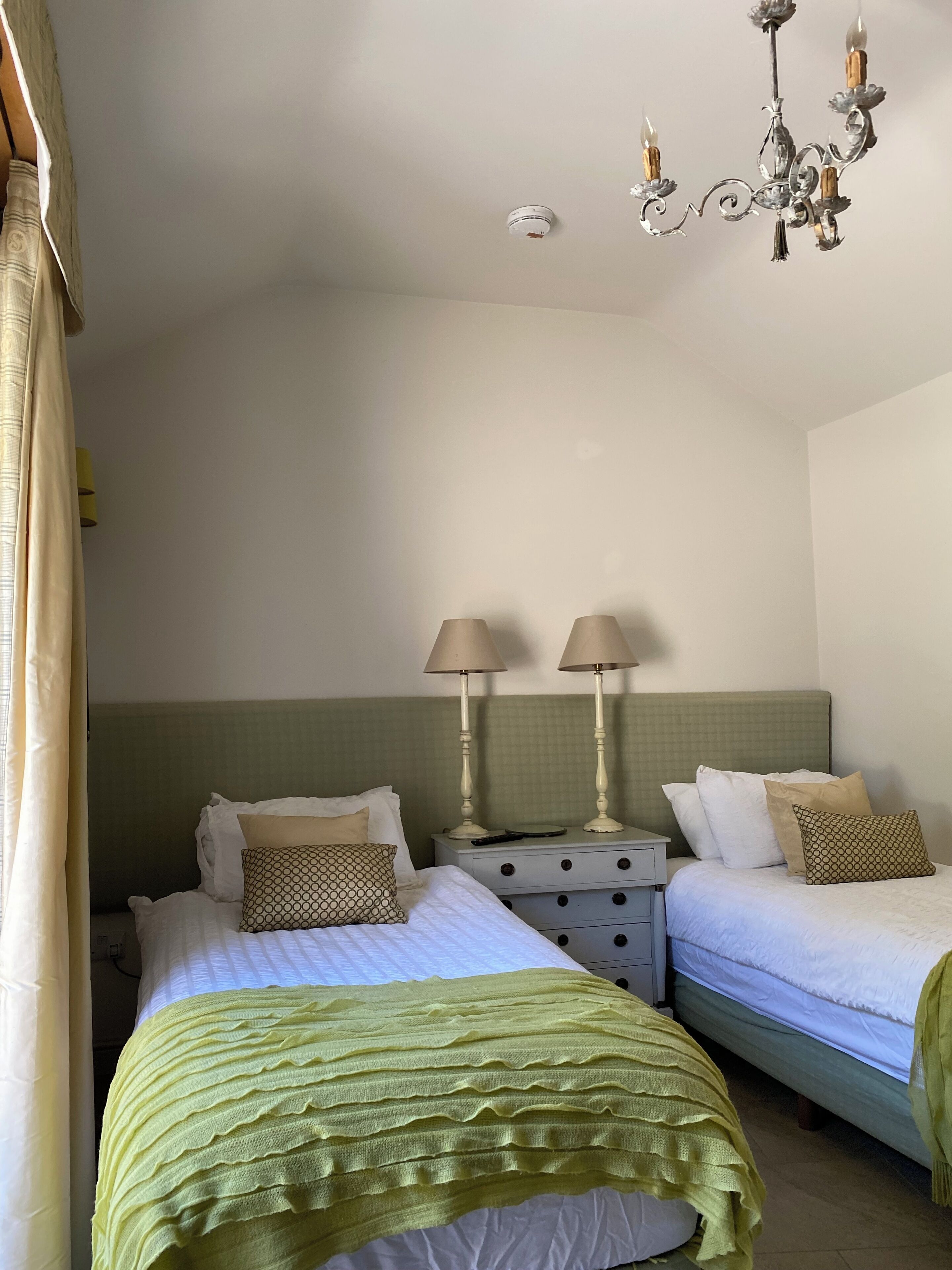 A hotel room with two beds, a green headboard, a chandelier, and a bedside table with lamps.