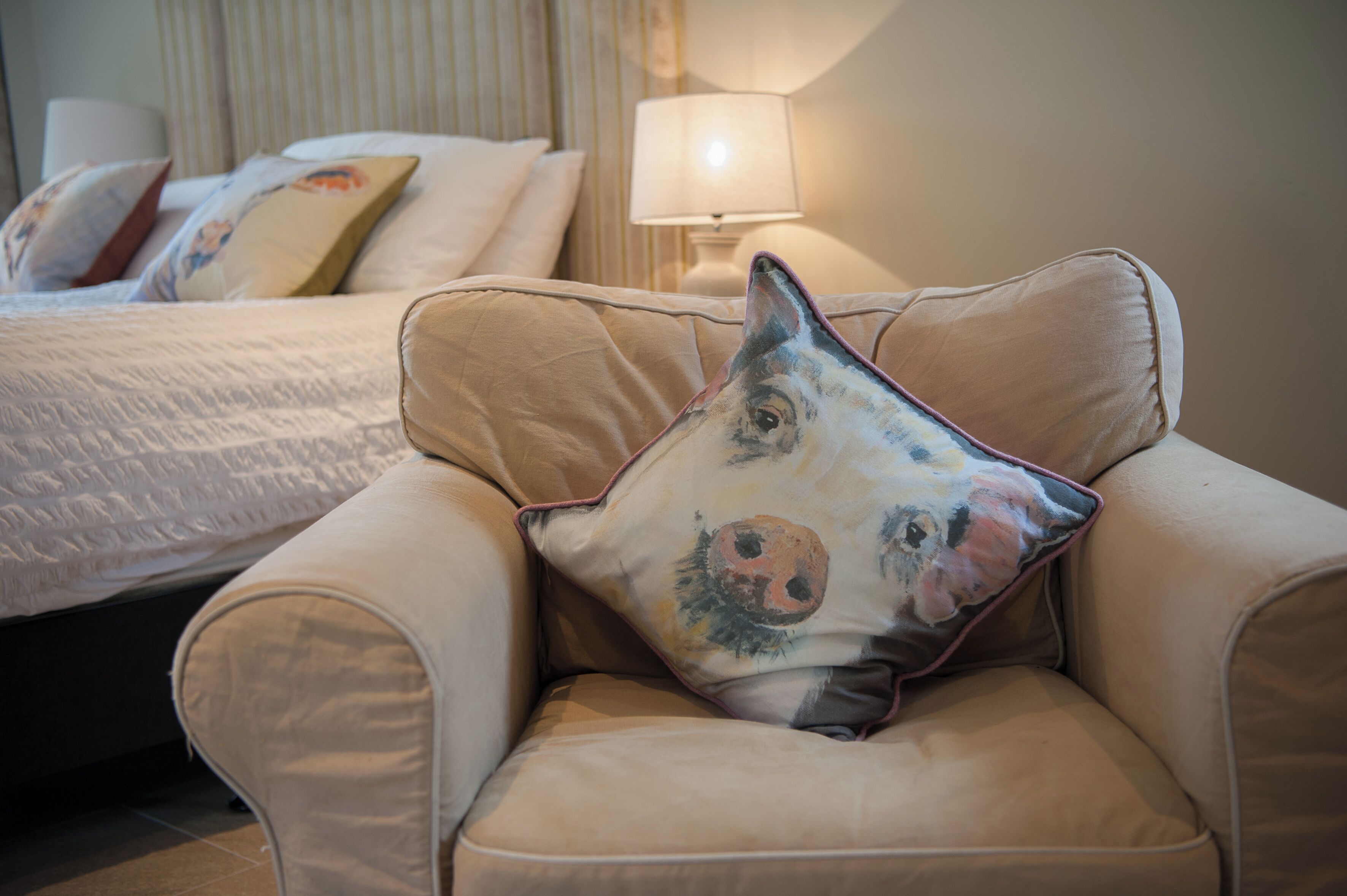 A hotel room with a bed, a lamp, and a chair with a pillow featuring a pig's face.