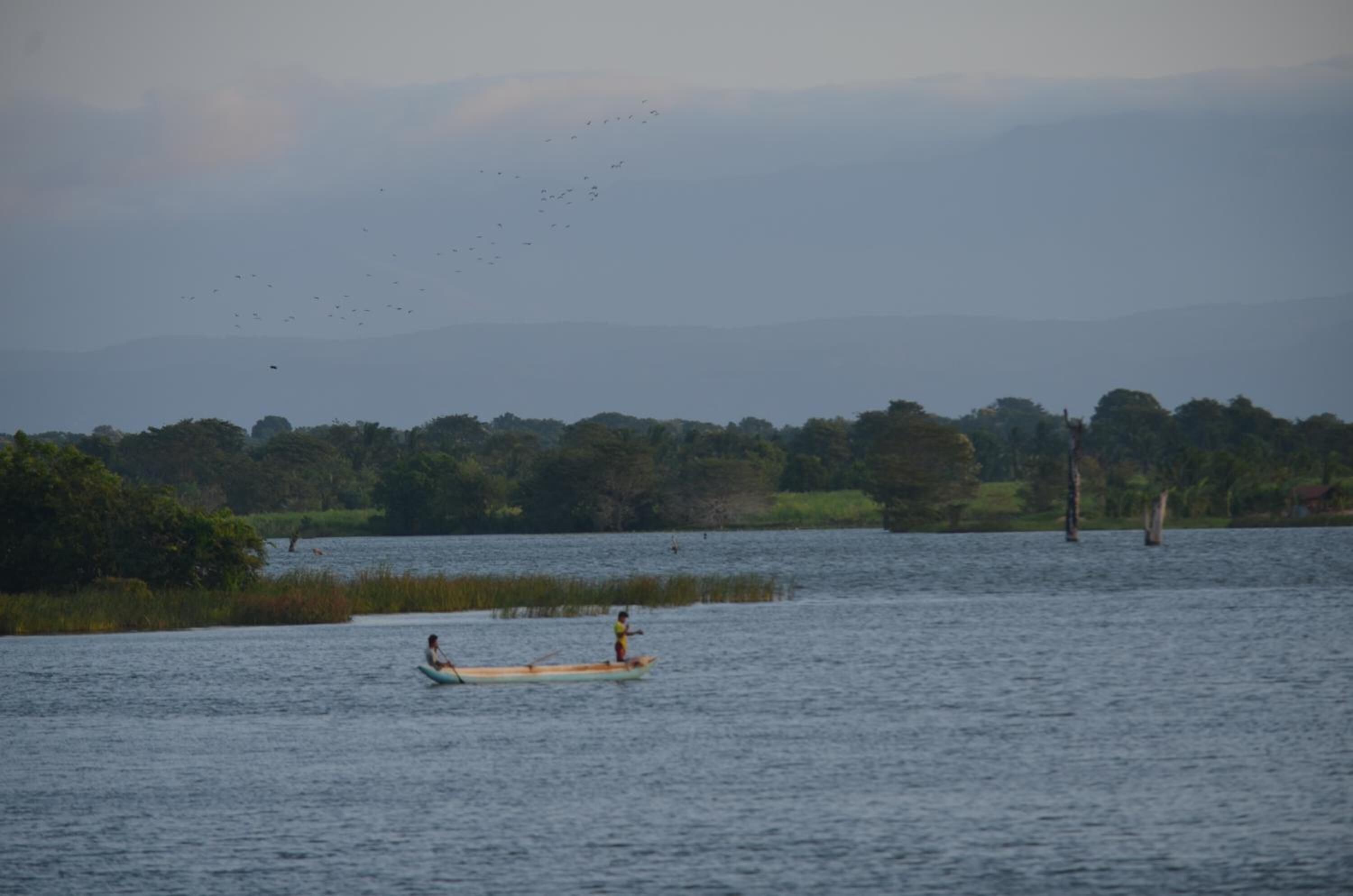 A calm lake with two canoes, one with a person paddling and another with a person standing.