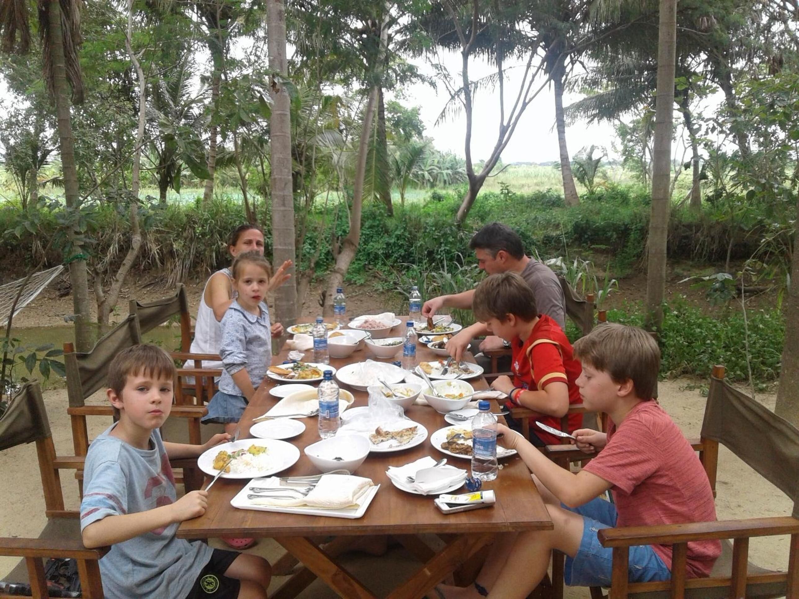 Un grupo de personas cenando en una mesa de madera al aire libre, rodeado de vegetación.
