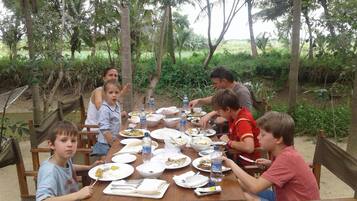 Un grupo de personas cenando en una mesa de madera al aire libre, rodeado de vegetación.