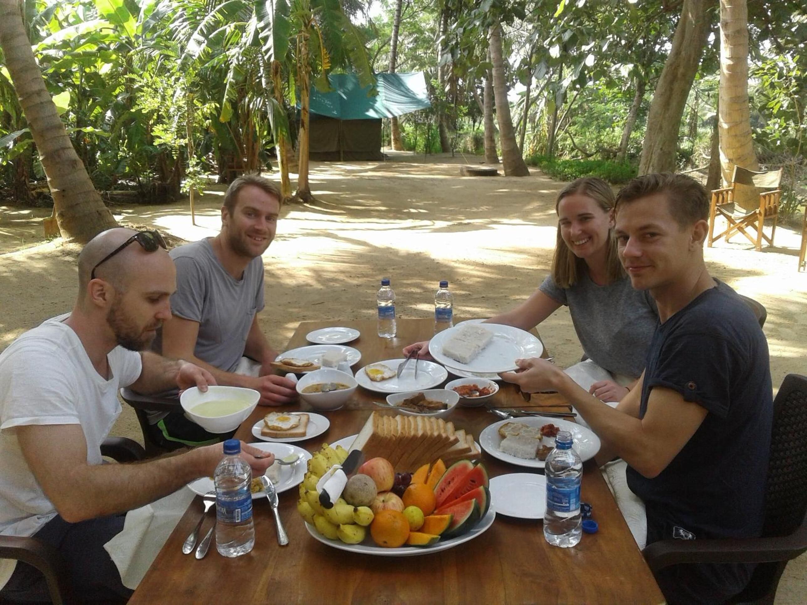 A group of people sitting around a wooden table outdoors, enjoying a meal with various dishes and fruits.