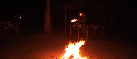 A campfire at night with a table and chairs in the background.