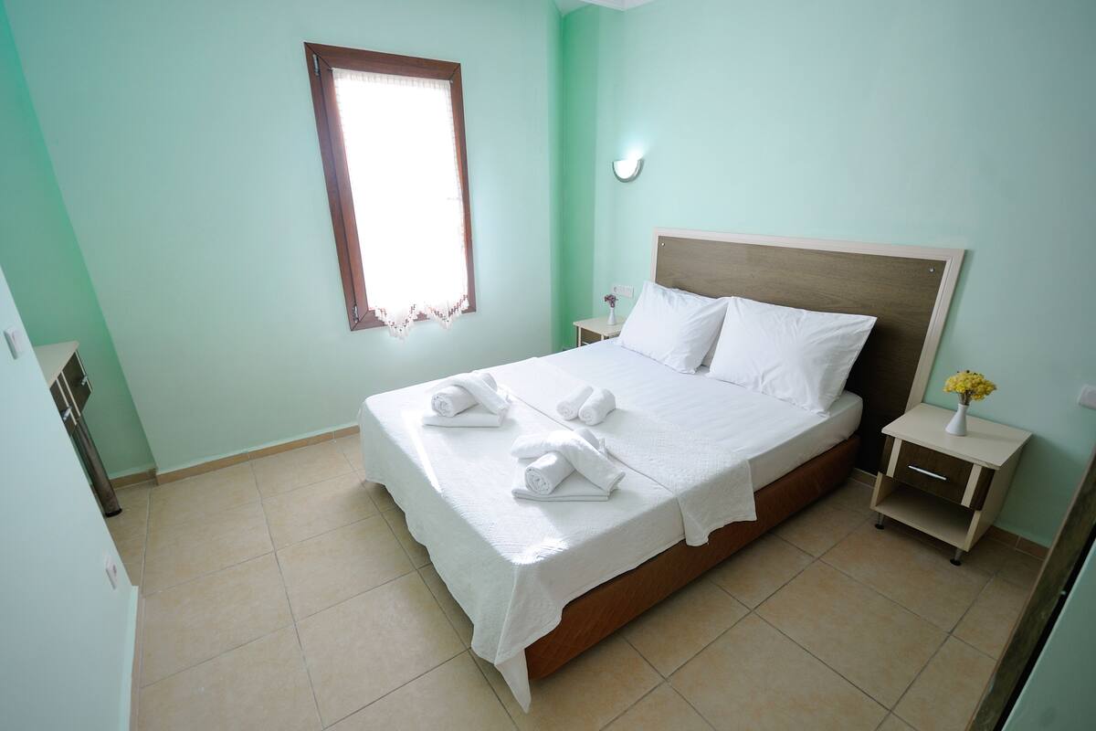 Una cama bien hecha con sábanas y toallas blancas, una mesita de noche con un florero, y una ventana con marco de madera.