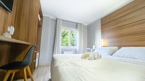 Ein Hotelzimmer mit einem Bett, einem Schreibtisch, einem Stuhl, einem Fenster mit Vorhängen und einem hölzernen Kopfteil.