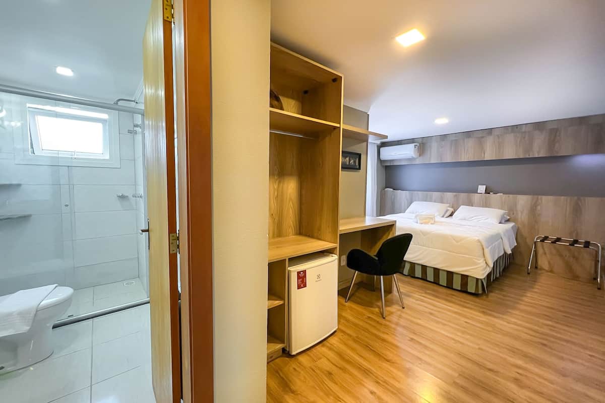 Une chambre d’hôtel de taille modeste, dotée d’un plancher en bois, d’un lit, d’un bureau, d’une chaise, d’un petit réfrigérateur et de toilettes.