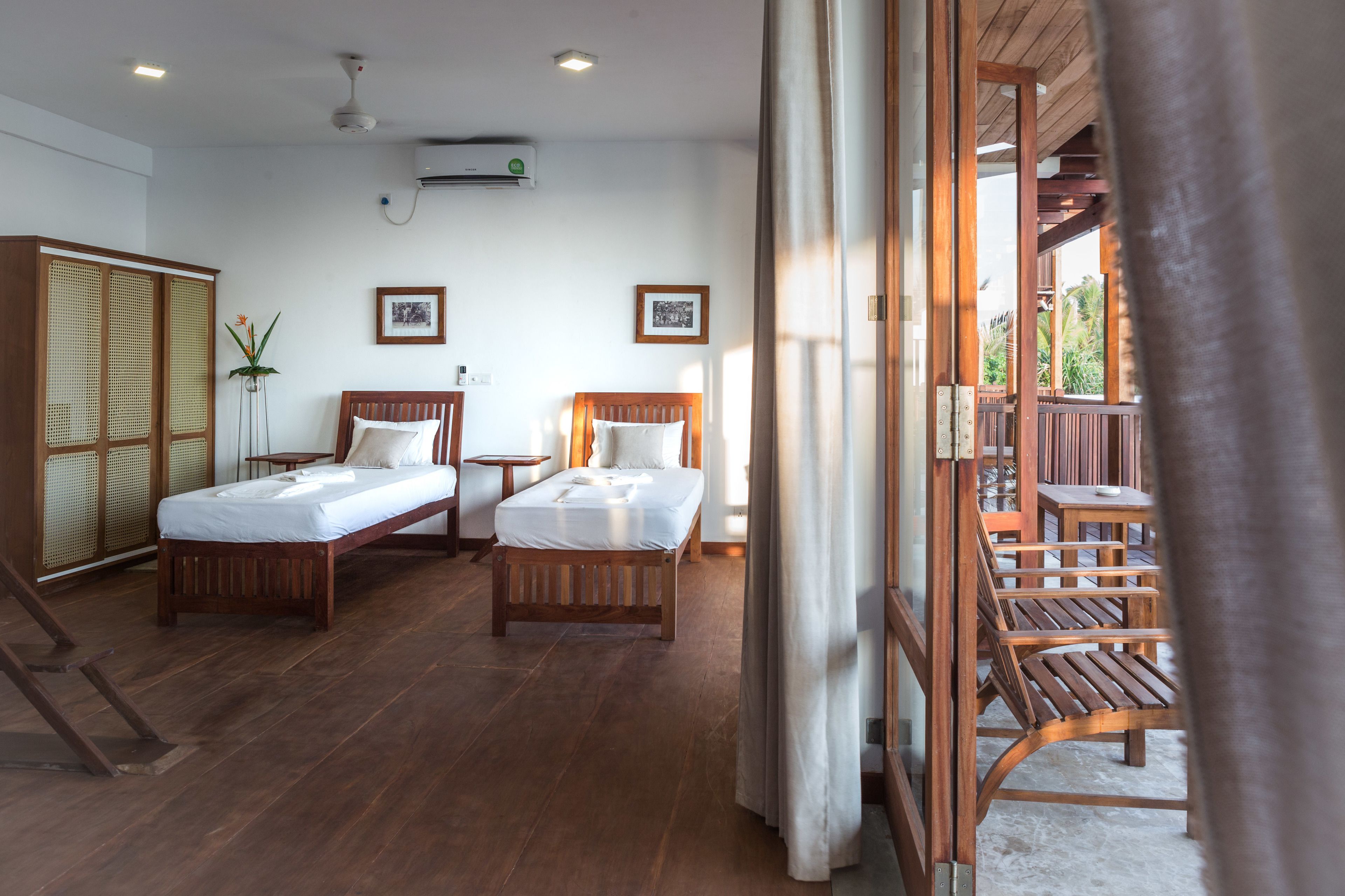 Une chambre d’hôtel avec deux lits, un plancher en bois, un balcon avec des chaises en bois et une vue sur la verdure.
