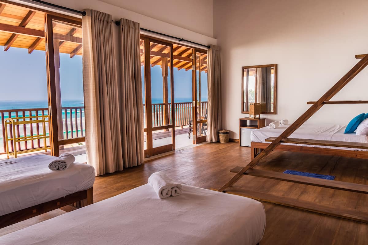 A hotel room with two beds, a wooden floor, a large window with a view of the ocean, and a balcony with a wooden railing.