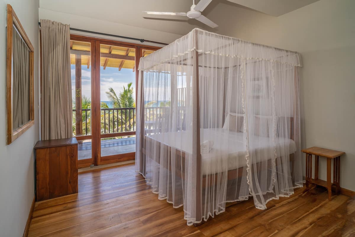 A room with a four-poster bed, wooden flooring, a ceiling fan, and a balcony with a view of palm trees.