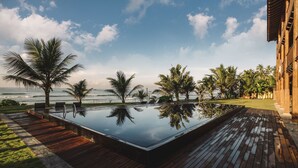Une piscine à débordement avec une terrasse en bois, des palmiers et une vue sur l’océan.
