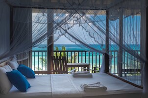 Een bed met witte lakens en blauwe kussens, een houten stoel, en uitzicht op de oceaan door transparante gordijnen.