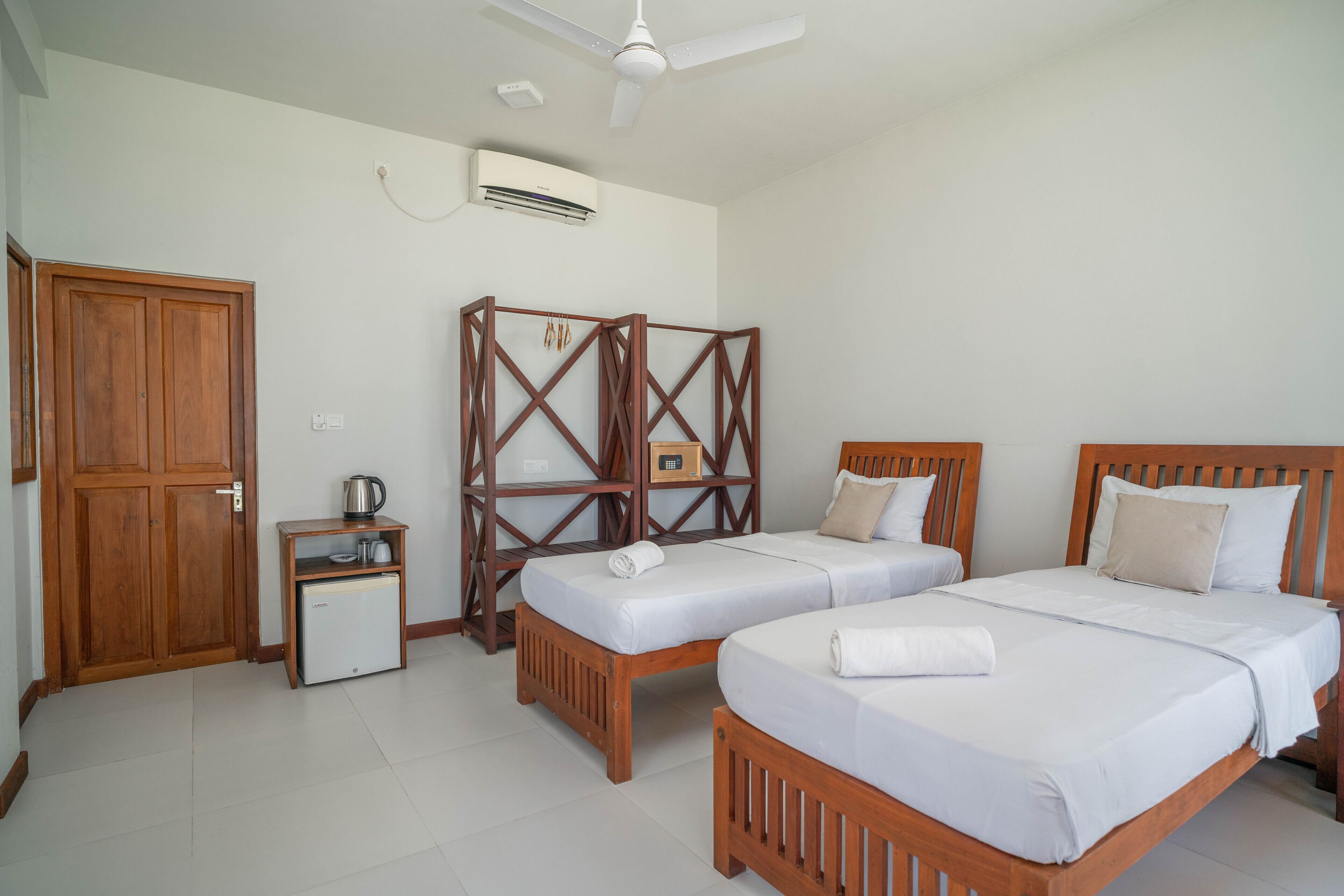 A hotel room with two wooden beds, a small kitchenette, a wardrobe, and a ceiling fan.