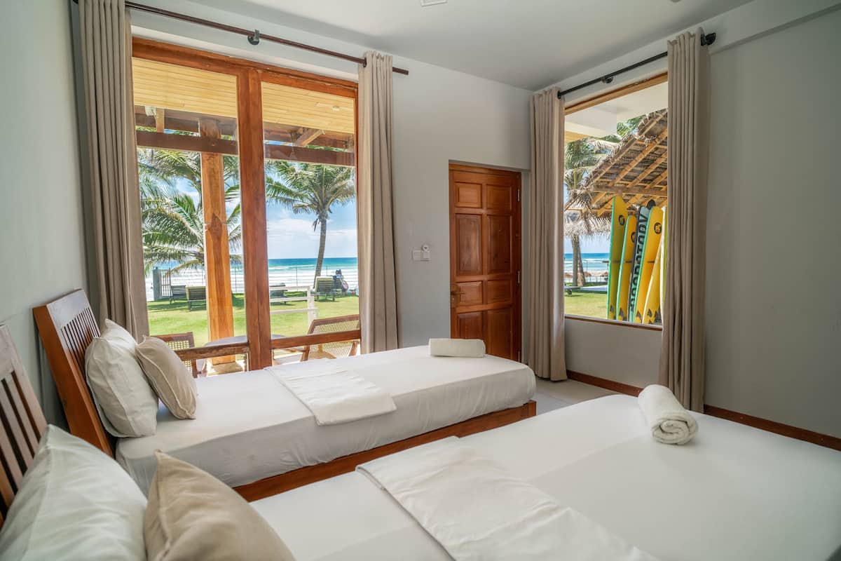 A hotel room with two beds, large windows with curtains, a wooden door, and a view of the beach and palm trees.