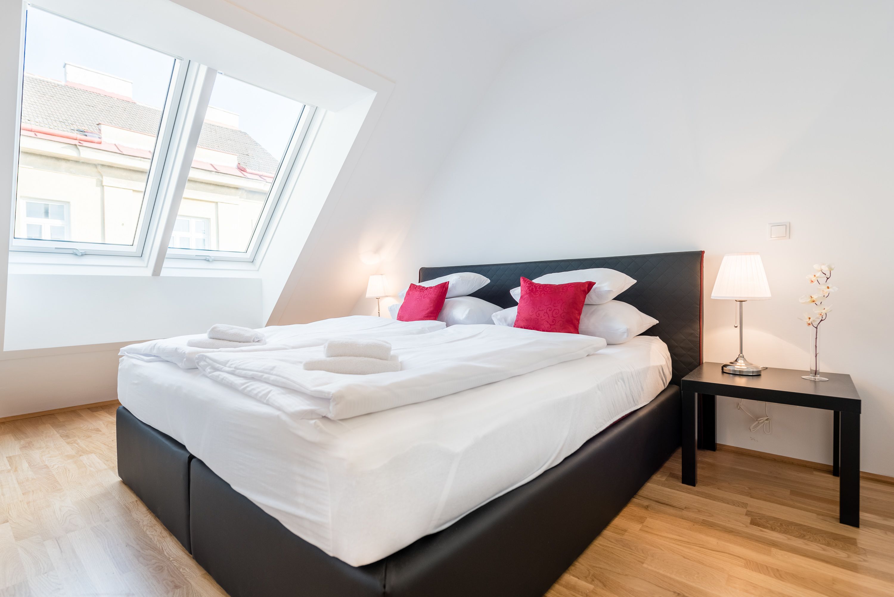 Un lit bien fait, avec du linge de lit blanc et des coussins rouges, une tête de lit noire et une table de chevet sur laquelle se trouvent une lampe et un vase de fleurs.