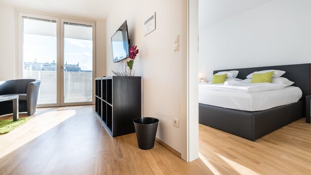 A modern hotel room with a bed, a desk, and a view of the city through large windows.