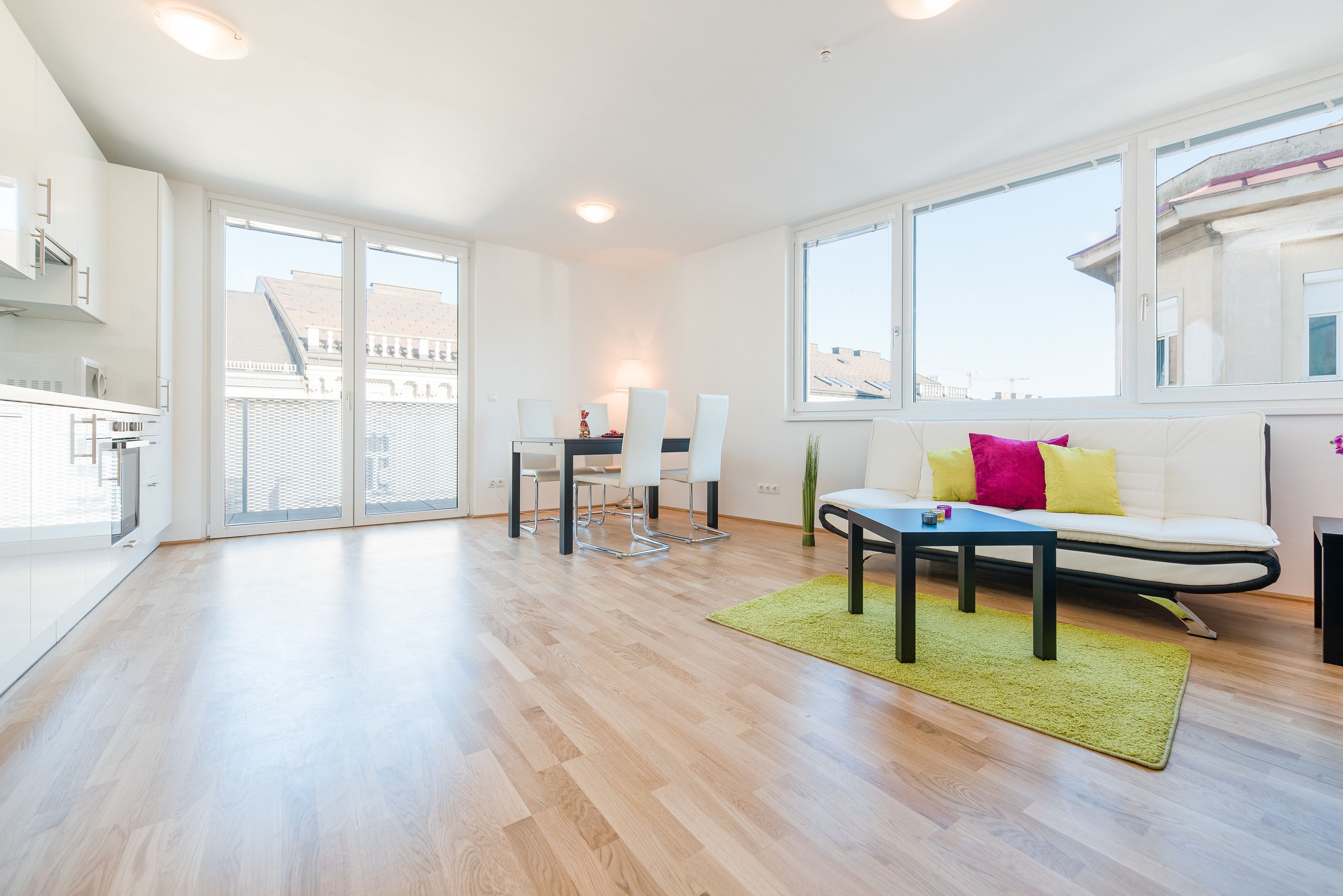 A modern living room with a sofa, dining table, and chairs. There is a large window providing a view of the city.
