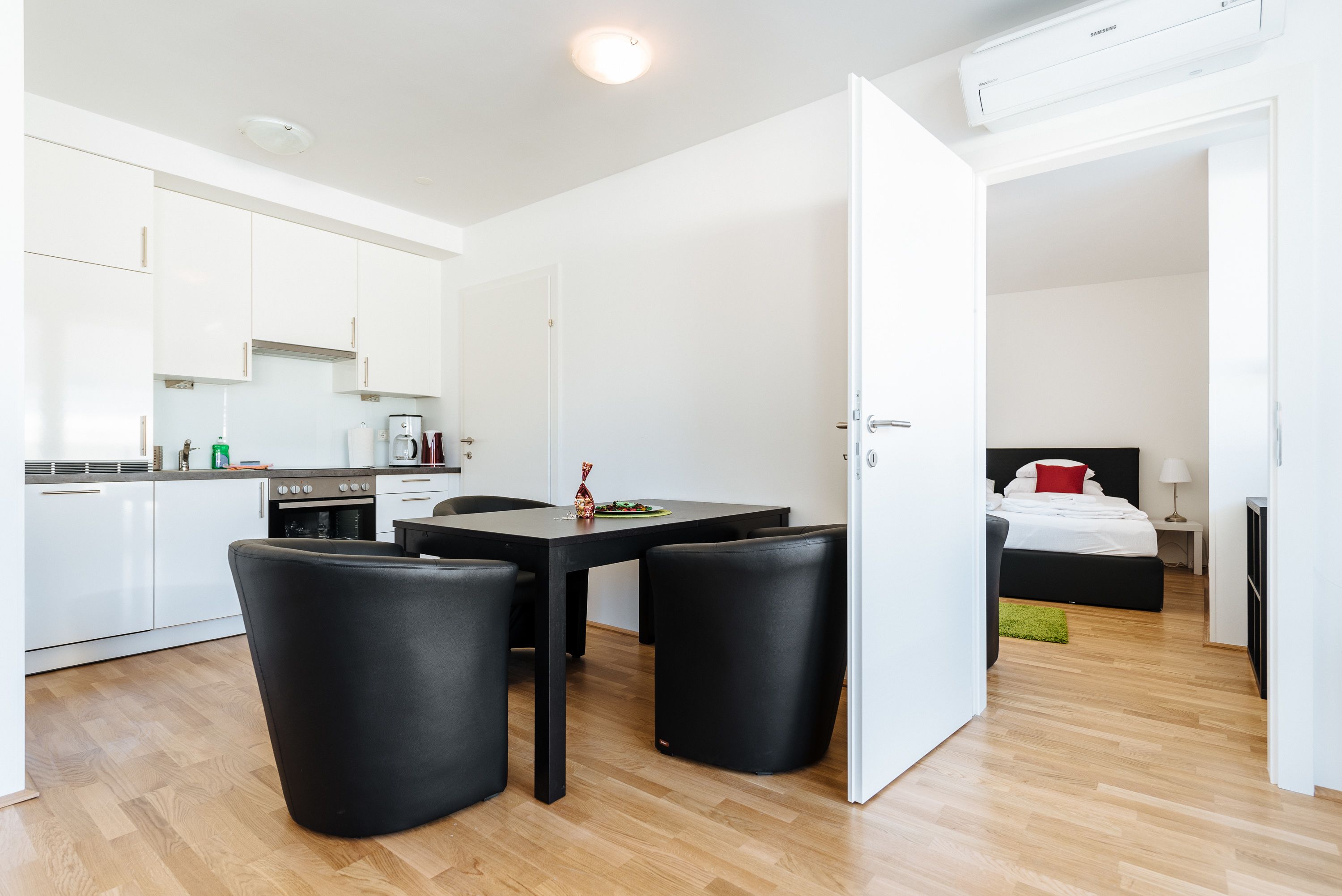 A modern, compact living space with a kitchenette, a dining area with black chairs, and a bedroom with a bed and nightstand.