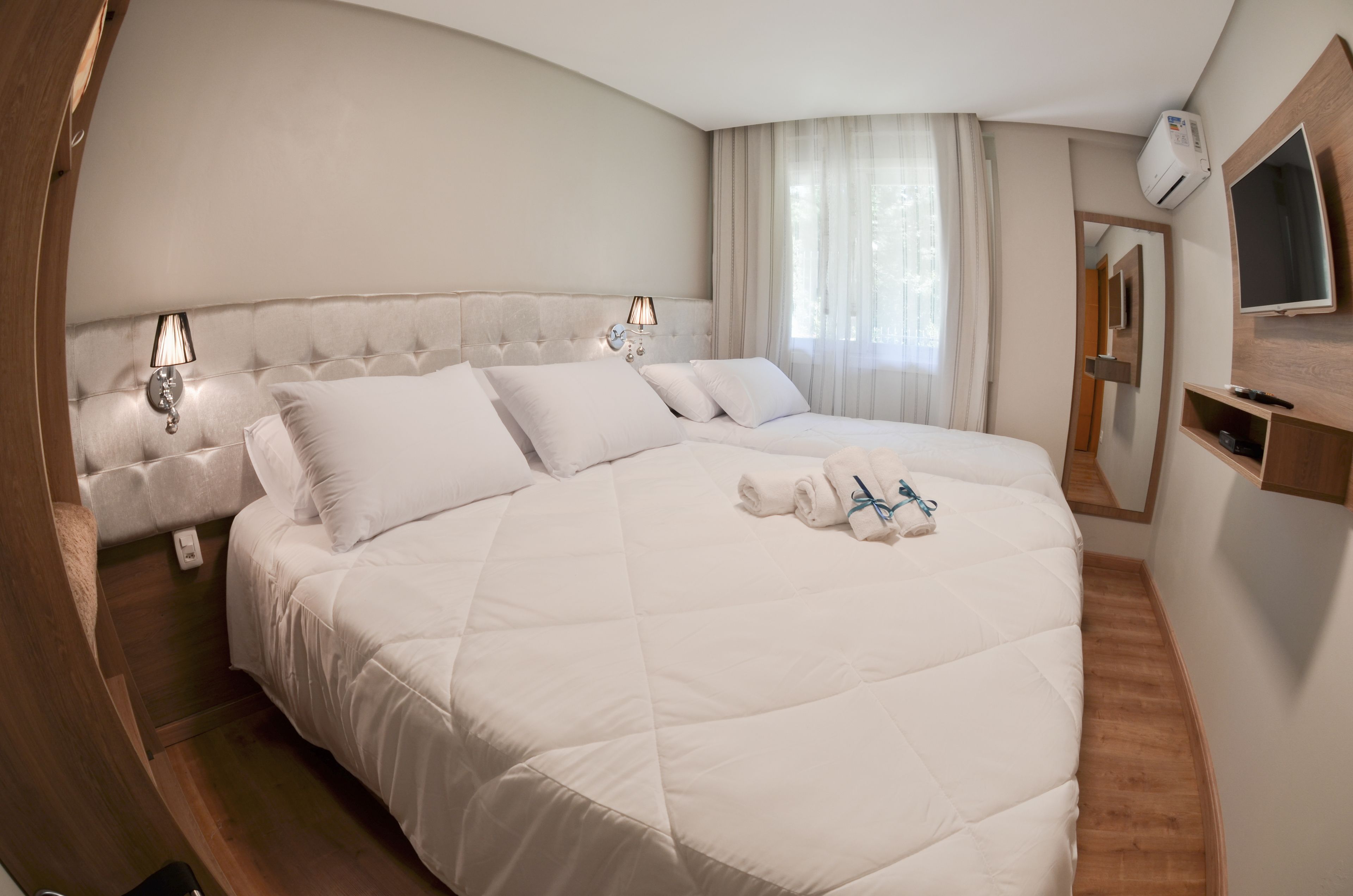 Una cama grande con ropa de cama blanca, dos lámparas de noche, un espejo y un televisor sobre una estantería de madera.