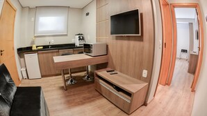 A compact kitchen with a microwave, coffee maker, and refrigerator. There is a flat-screen TV mounted on the wall and a small seating area with a couch. - Viena Flats (Gramado)