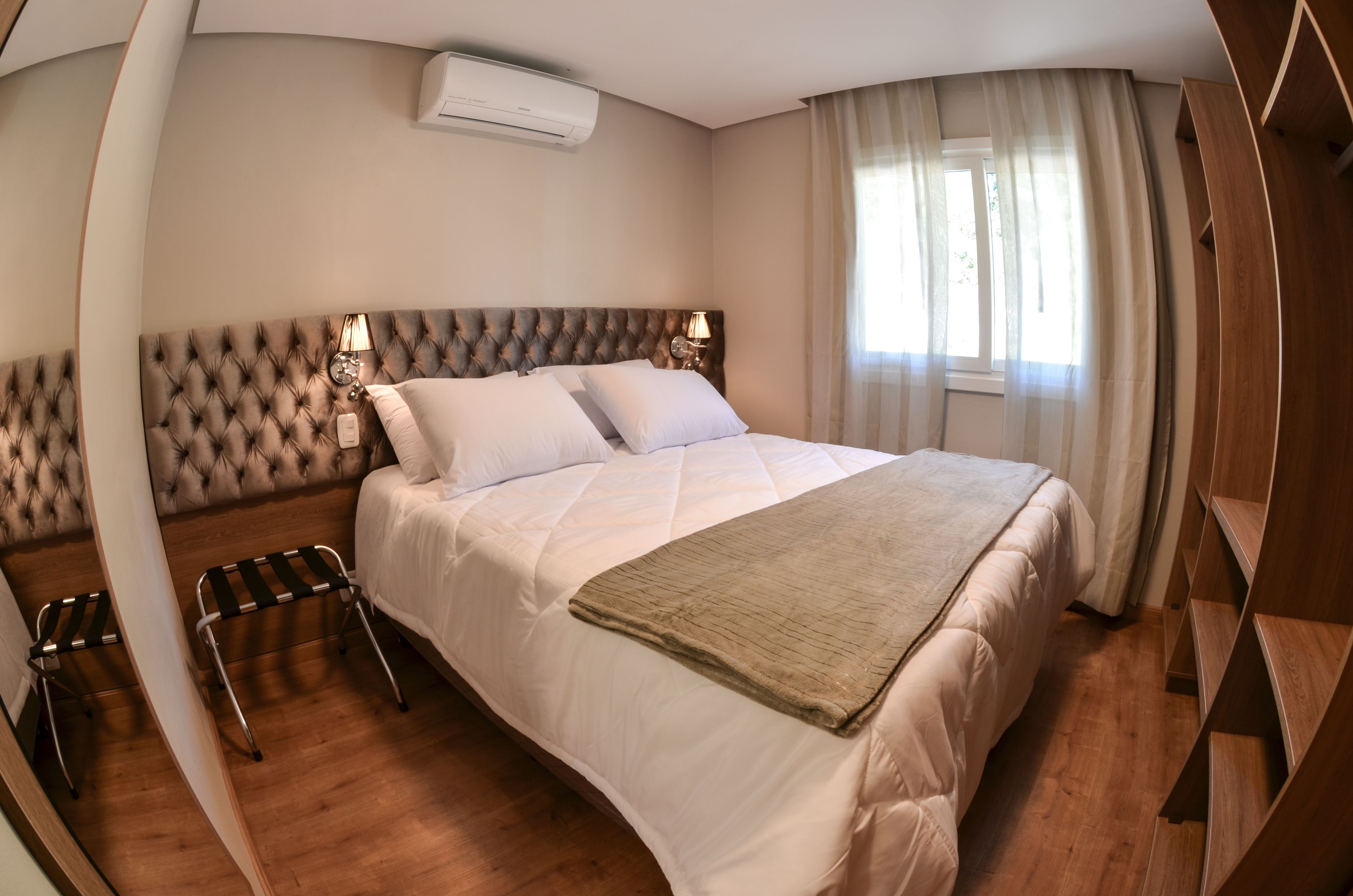Una cama bien hecha con cabecera capitonada, una lámpara de noche y una ventana con cortinas.