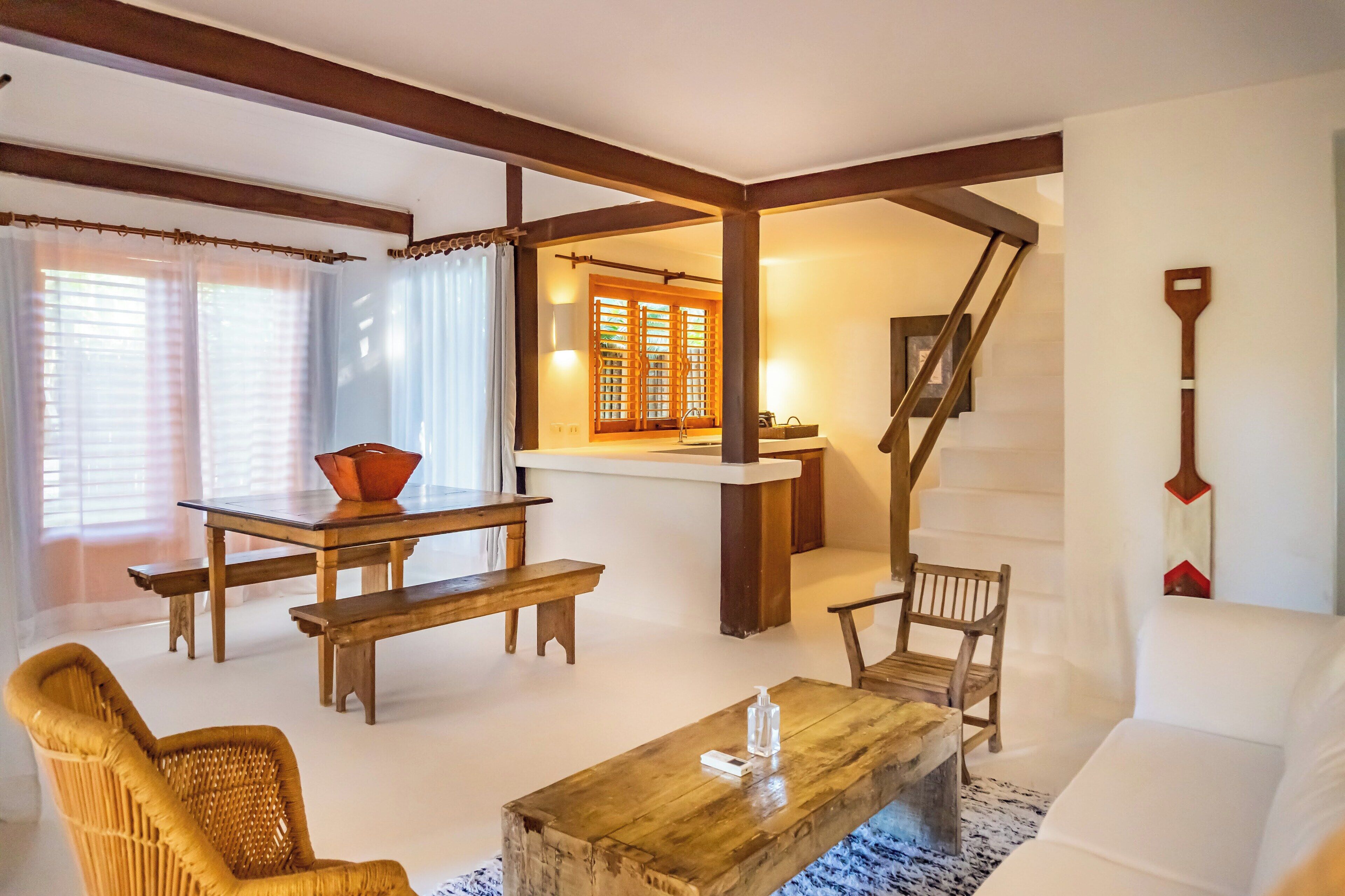 Una sala de estar acogedora con un banco de madera, un sillón de mimbre, una mesa de centro y un sofá blanco. Una escalera conduce a un nivel superior con barandilla de madera.