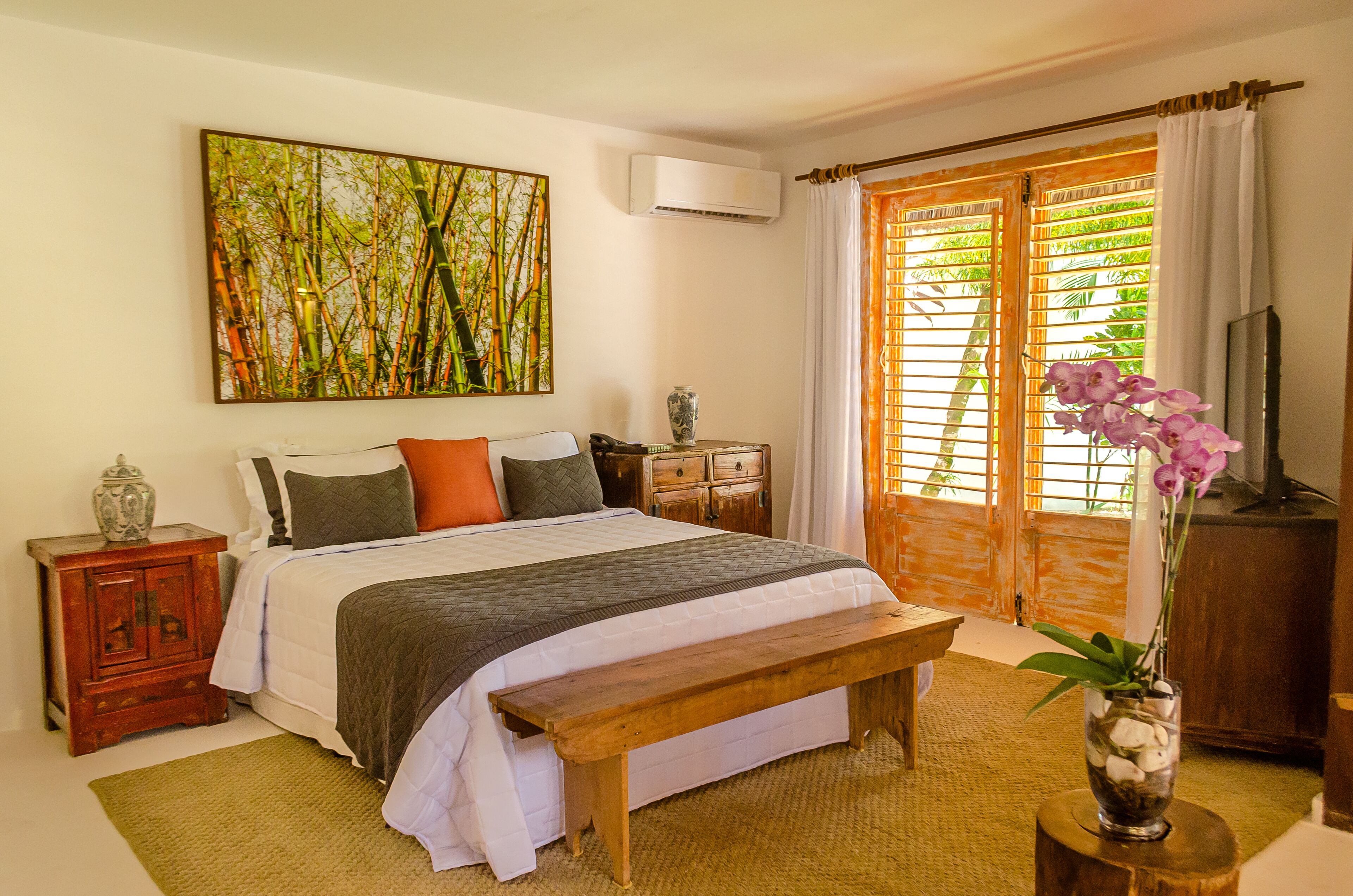 Une chambre à coucher avec un lit, un banc en bois, une table de chevet, un vase avec des fleurs et un tableau accroché au mur.