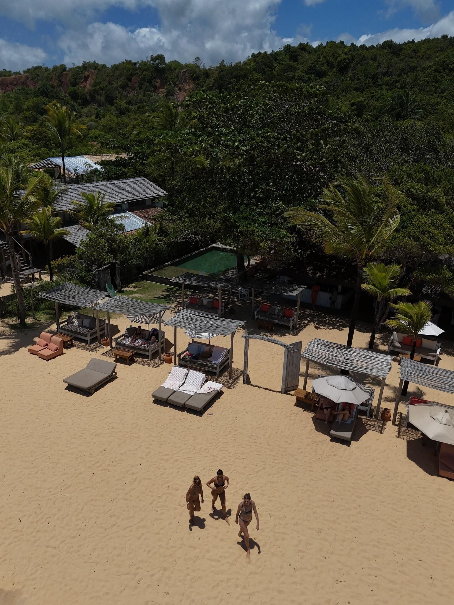 A beach with sun loungers and umbrellas, a pool, and a forested hill in the background.