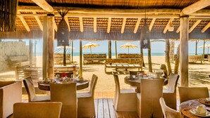 Un área de comedor al aire libre con mesas y sillas, sombrillas y vista a la playa.
