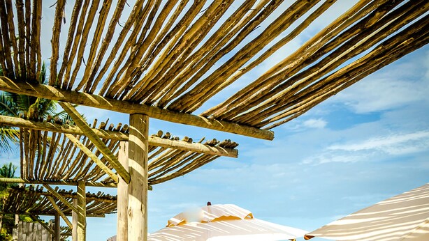Un área de descanso frente a la playa con mobiliario de madera, sombrillas y vista al mar.