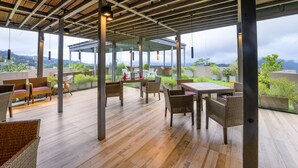 Un patio al aire libre con piso de madera, mobiliario de mimbre y ventanas amplias que ofrecen vistas a zonas verdes y edificios lejanos.