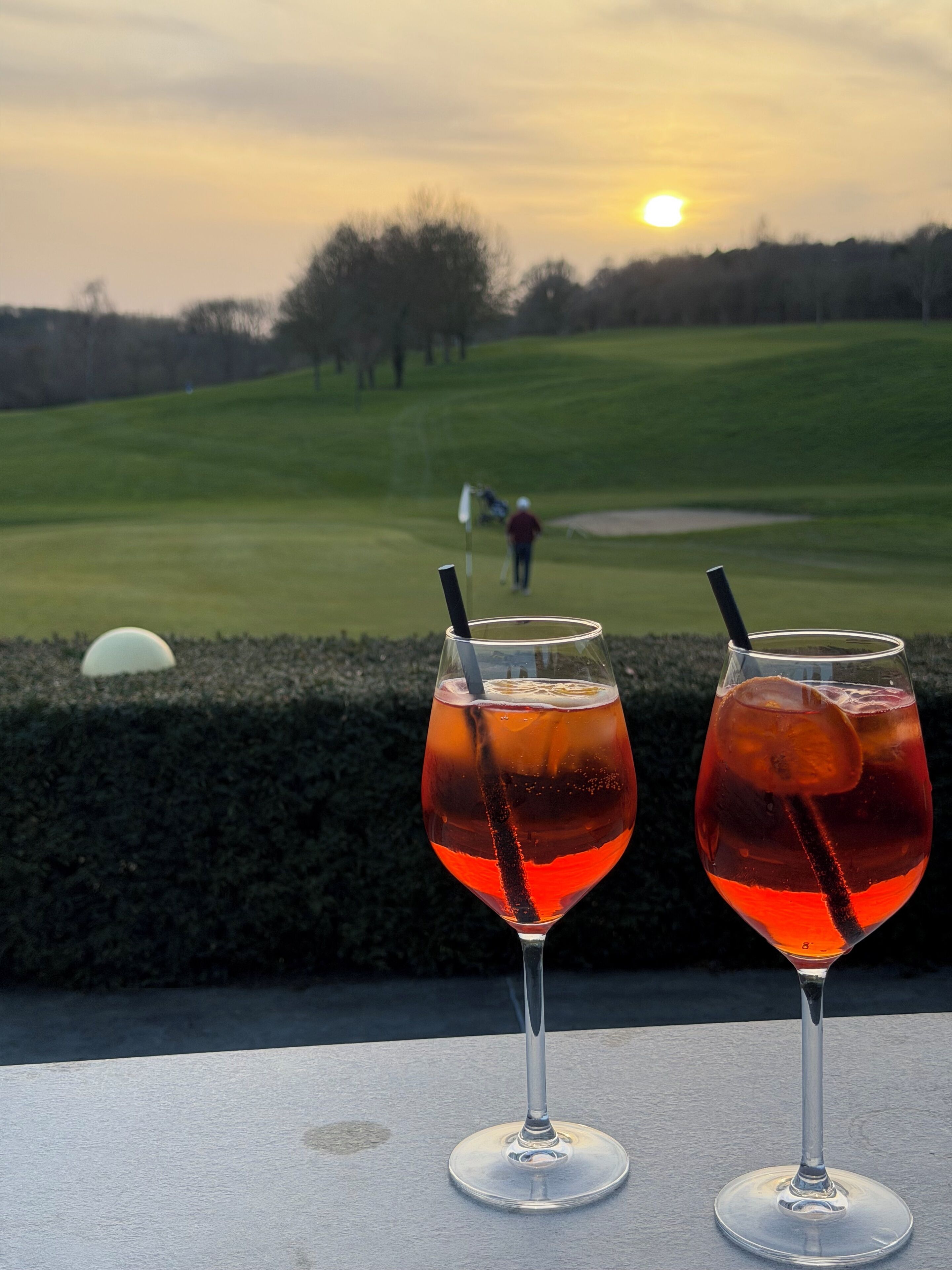 Two glasses of red beverage on a table with a golf course and sunset in the background.
