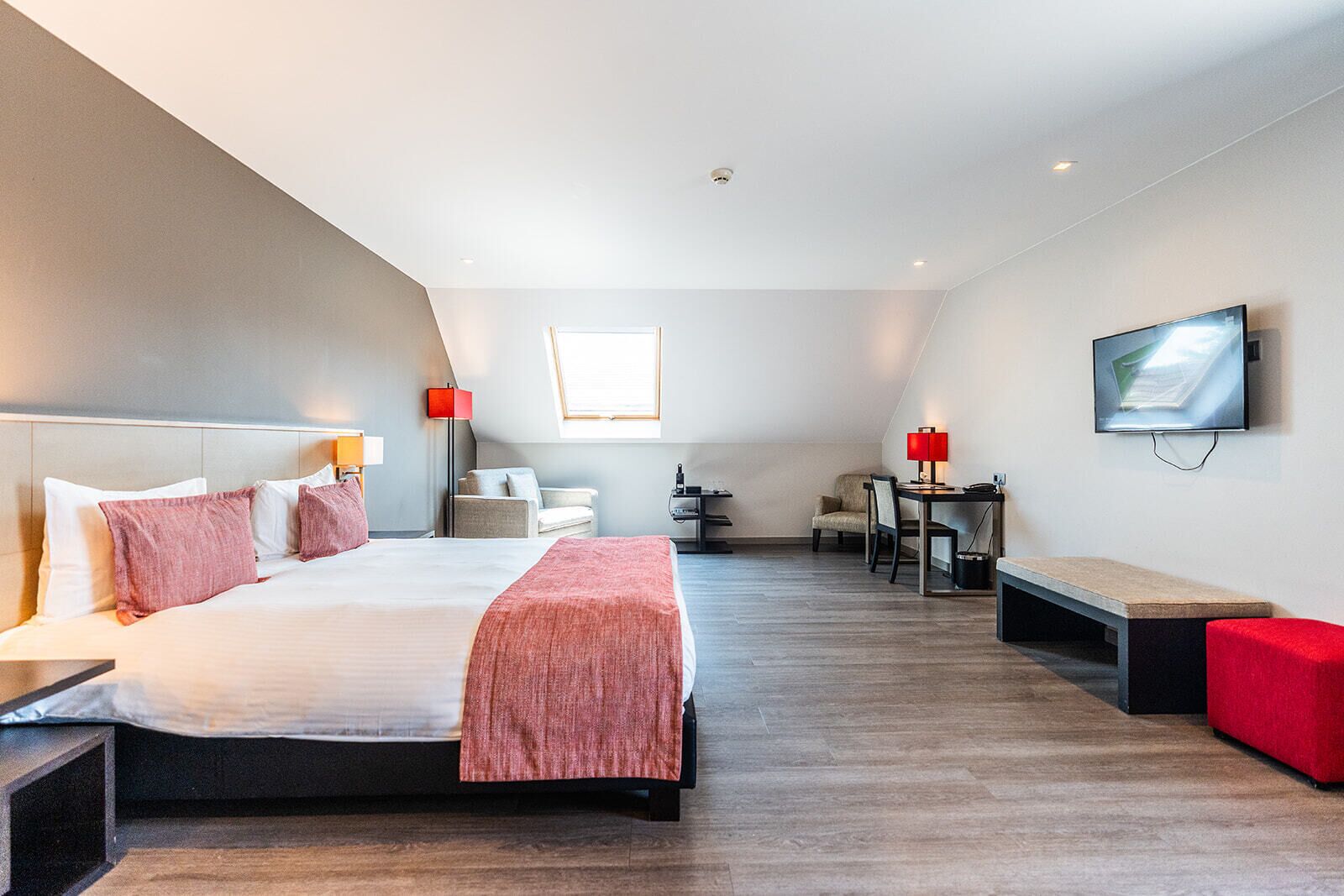 A modern hotel room with a large bed, a desk, a chair, a TV, and a small table.