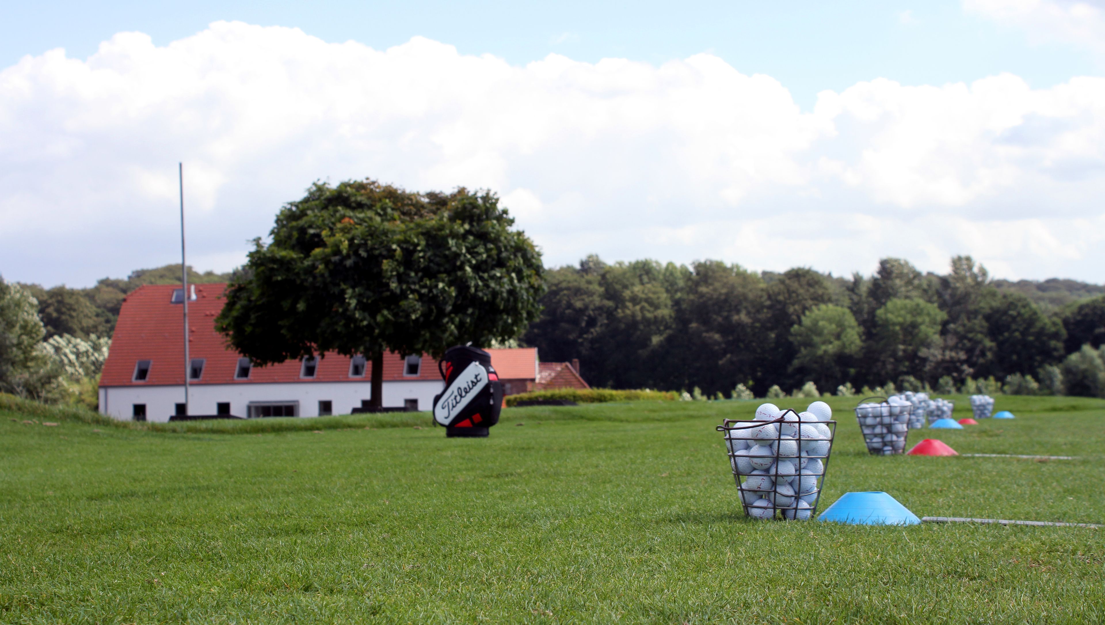 Un terrain de golf avec des balles de golf soigneusement disposées dans des paniers, un bâtiment au toit rouge et un grand arbre en arrière-plan.