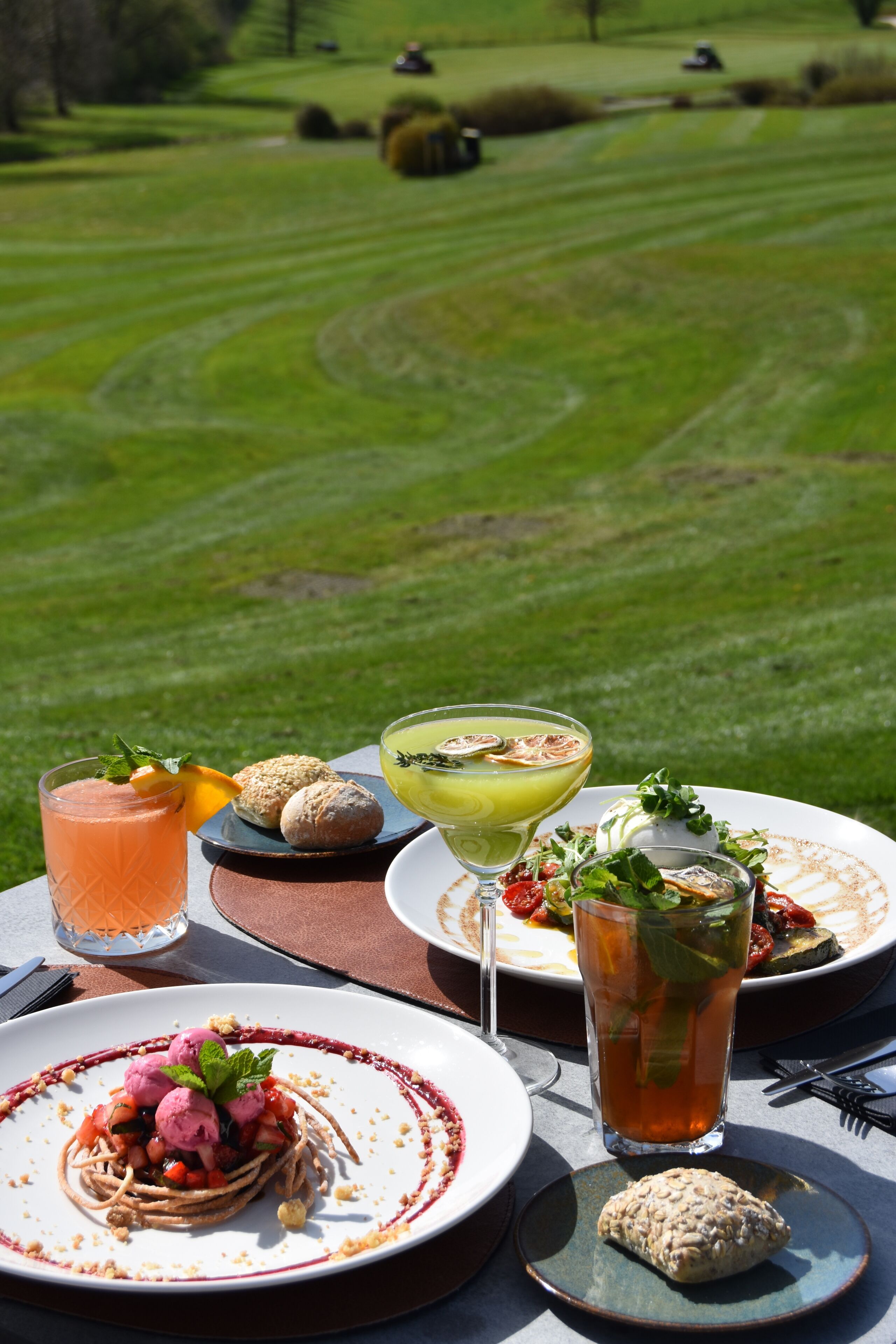 A table with a meal and drinks, set against a backdrop of a well-manicured lawn and distant trees.