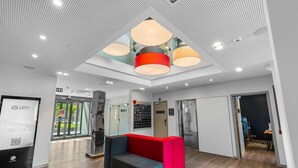 Una zona de recepción moderna con un área de descanso en rojo y azul, una entrada con puerta de cristal y un panel informativo digital.