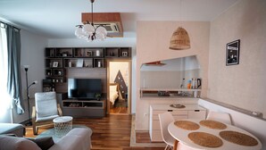 A modern living room with a sofa, TV, and entertainment unit; a dining area with a table and chairs; and a kitchen with a counter and appliances.