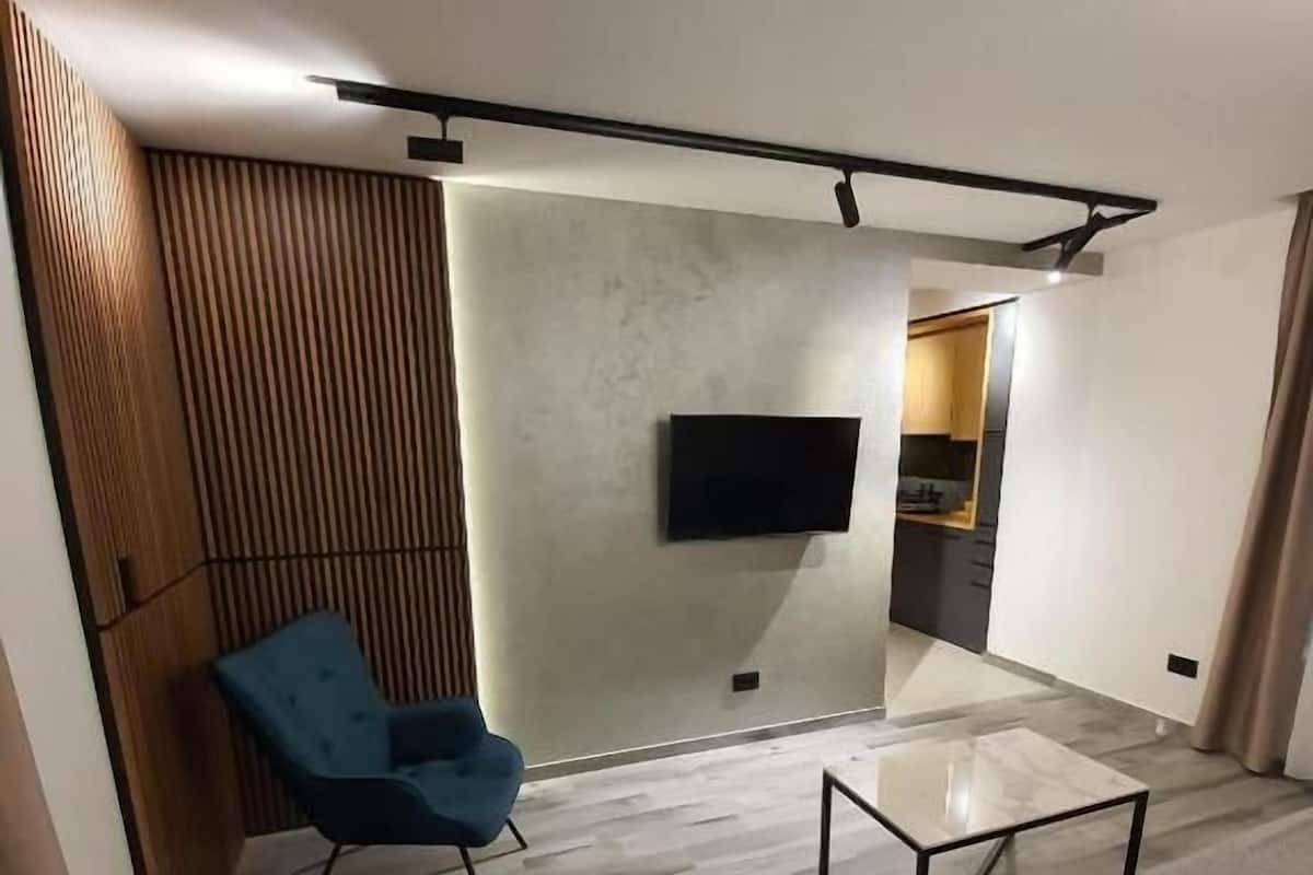 A modern living room with a sofa, a blue chair, a glass coffee table, and a flat-screen TV mounted on the wall.