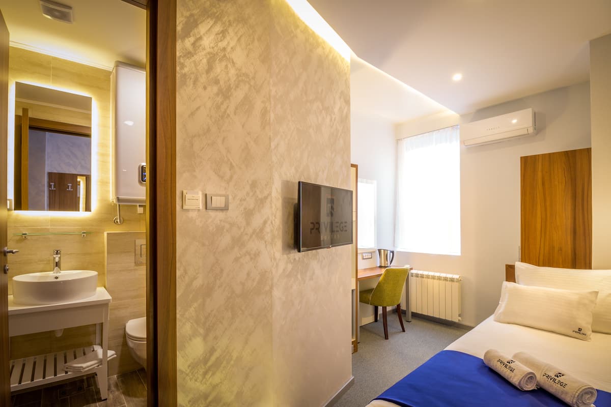 A hotel room with a bed, a desk with a chair, a TV, a bathroom with a sink and toilet, and a window.