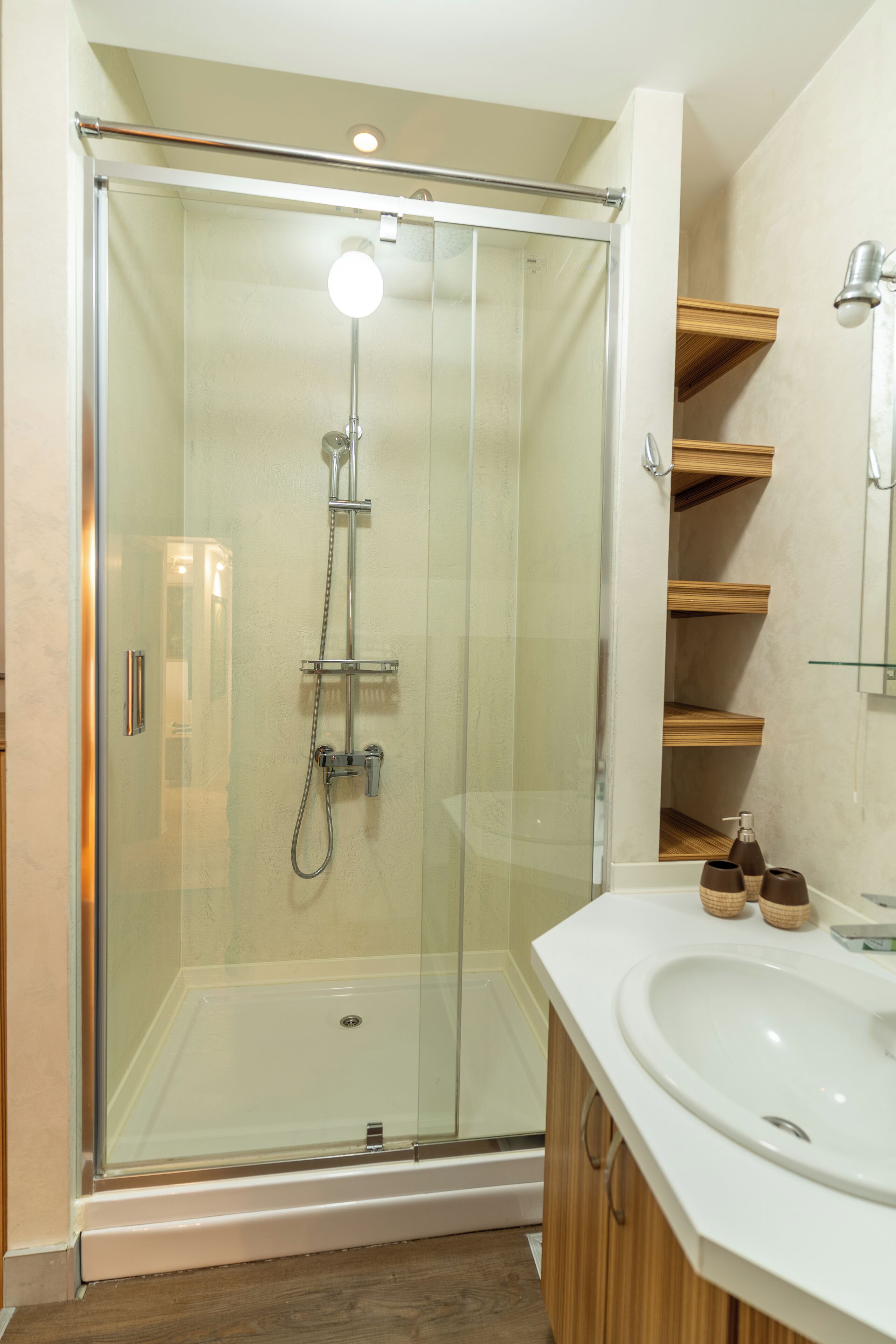 Una ducha con cerramiento de vidrio, un cabezal de ducha de mano, un lavabo blanco con un espejo encima y estantes de madera sobre el lavabo.