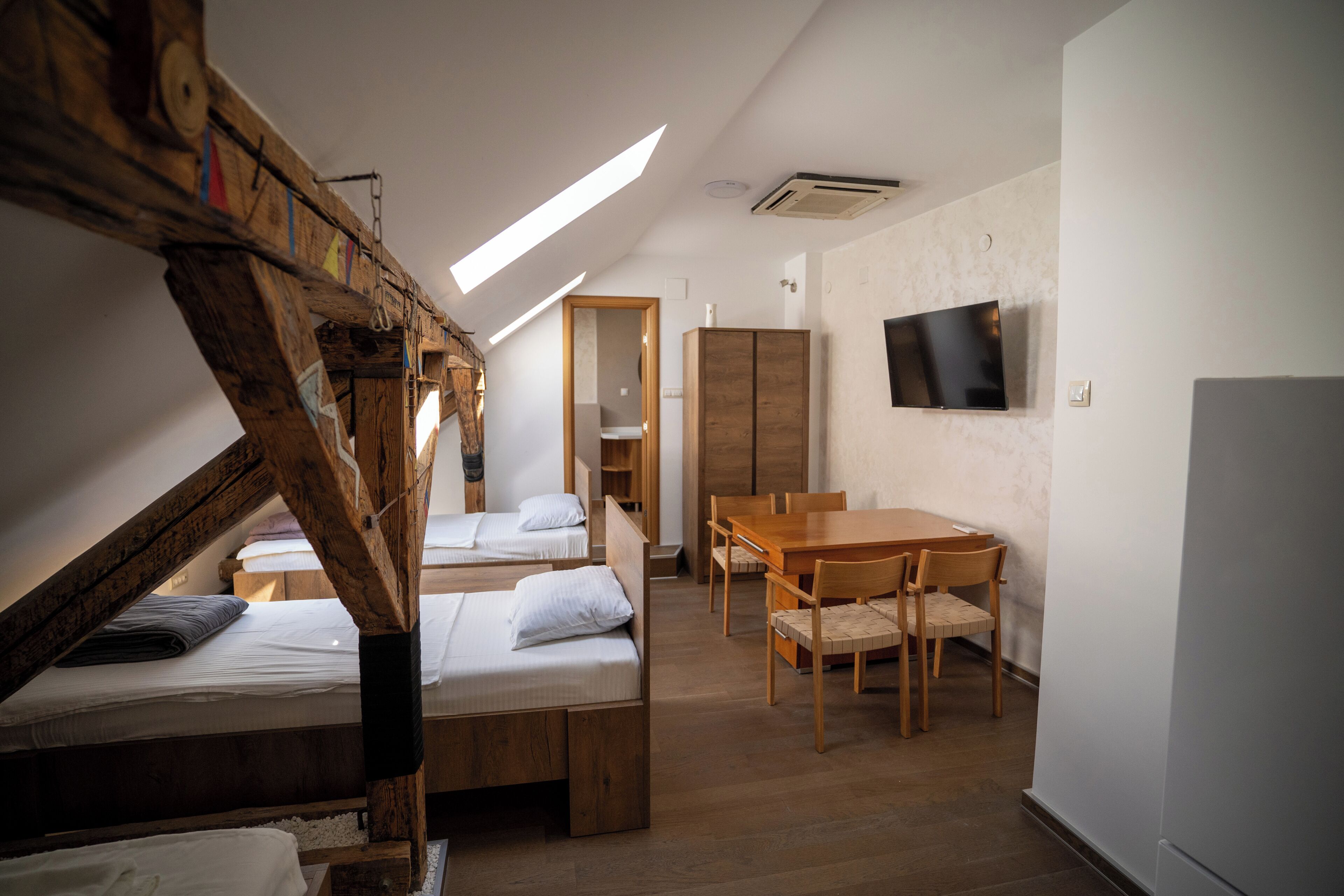 A loft-style bedroom with a wooden staircase, two beds, a dining table, and a TV mounted on the wall.