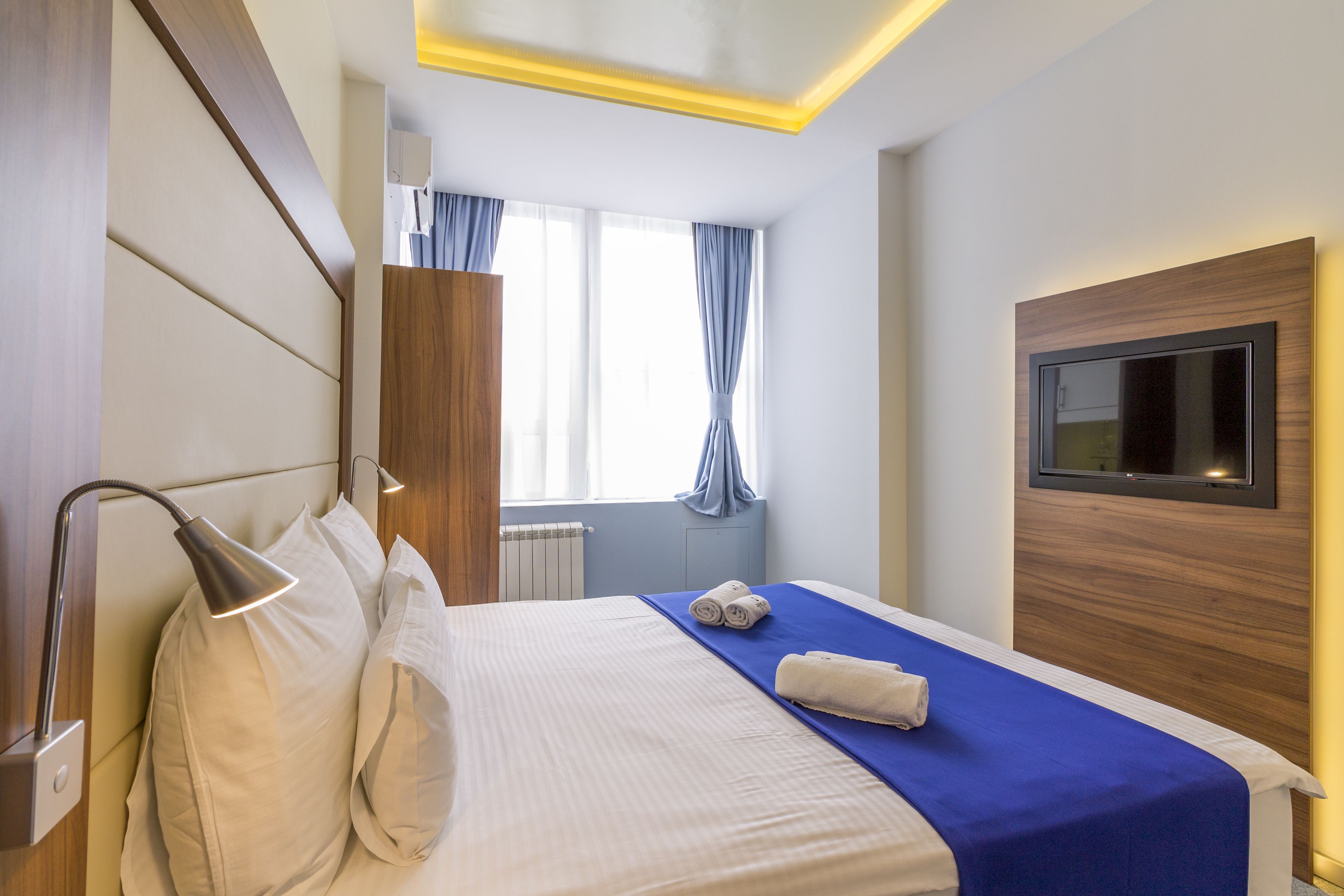 Ein Hotelzimmer mit Bett, Wandfernseher, Fenster mit Vorhängen und einer Nachttischlampe.