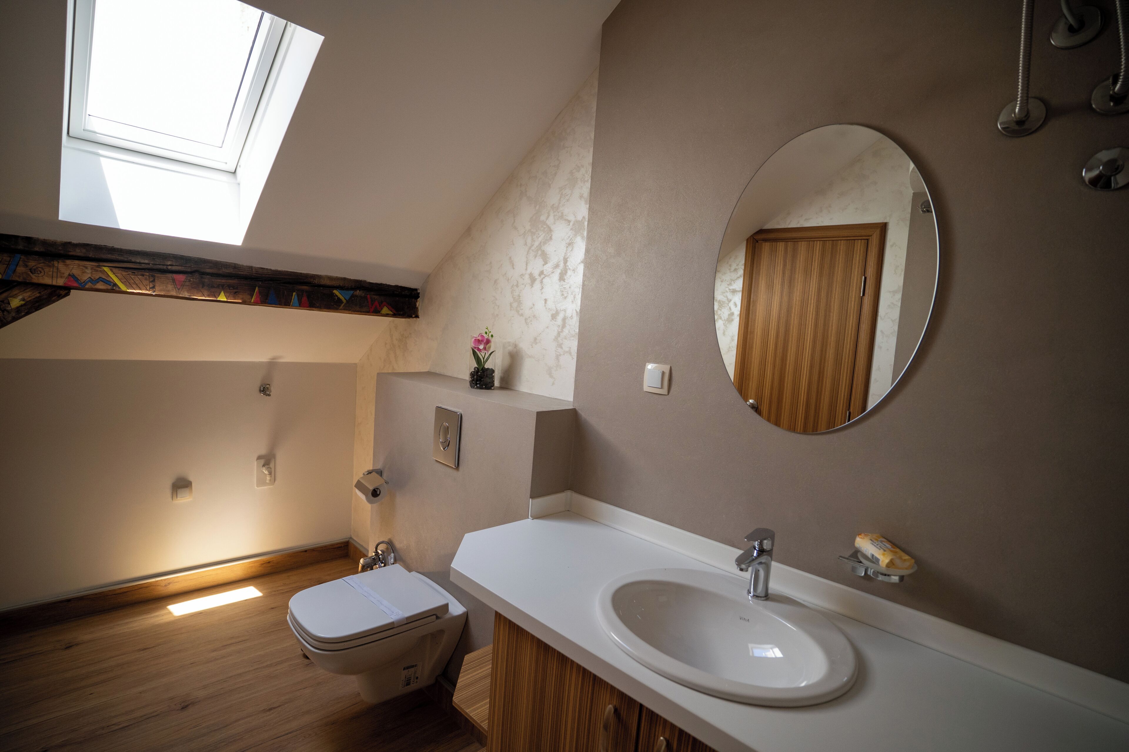 A bathroom with a skylight, a round mirror, a sink, and a toilet.