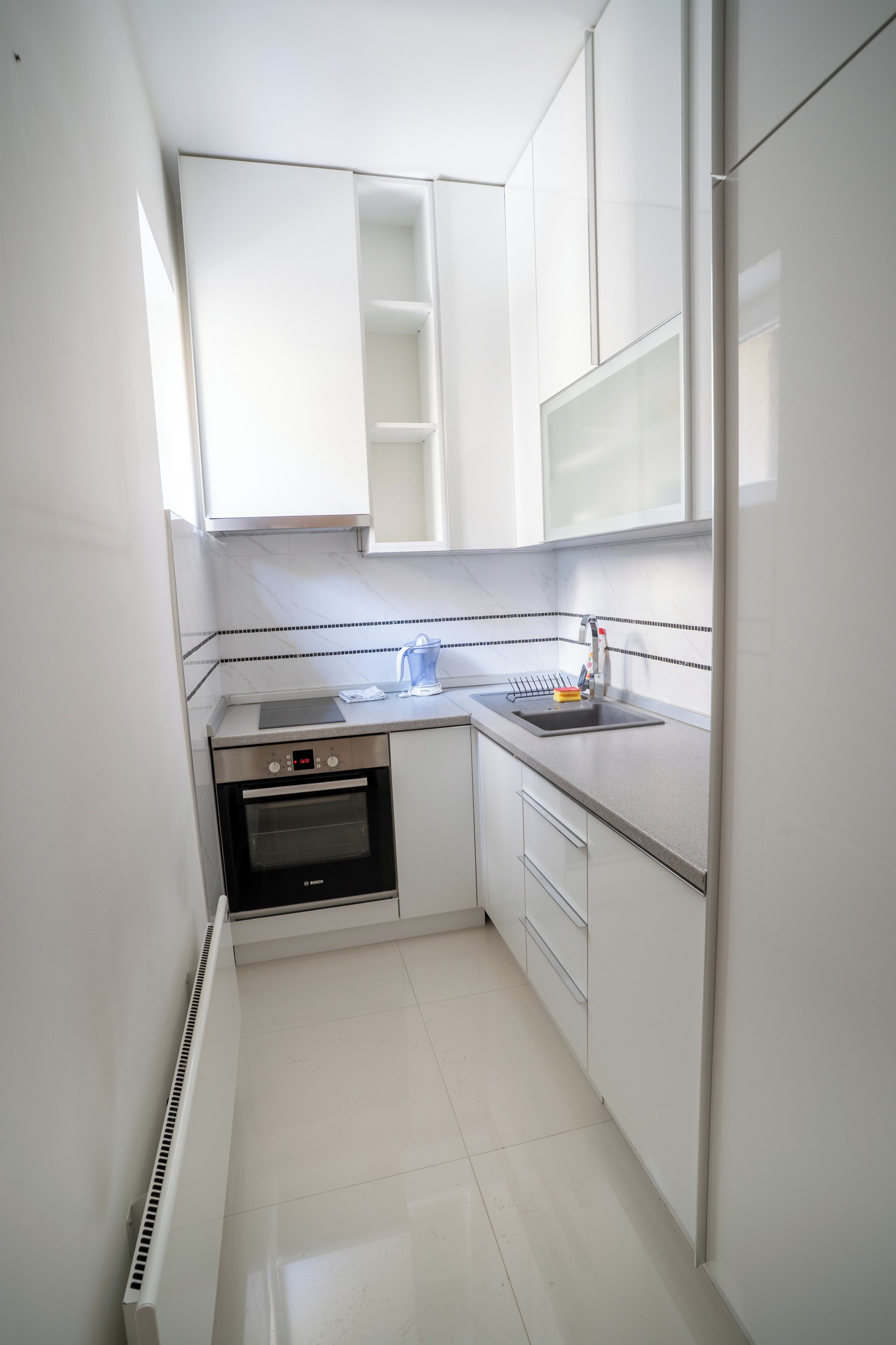 Cocina compacta con armarios blancos, horno integrado, lavamanos y ventana sobre la encimera.