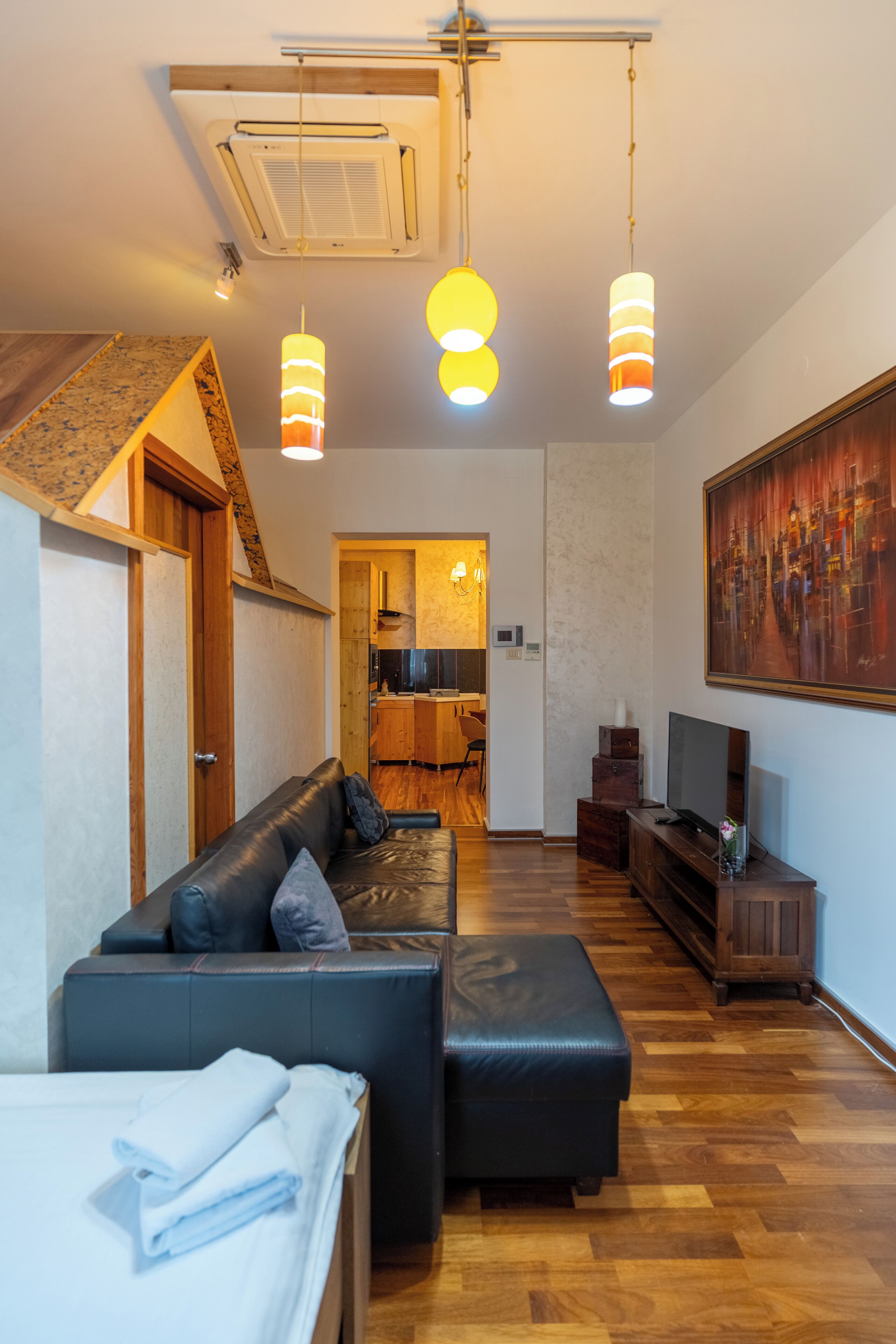 Una sala de estar moderna con sofá, reposapiés y televisor de pantalla plana. Tiene piso de madera, ventilador de techo e iluminación colgante.