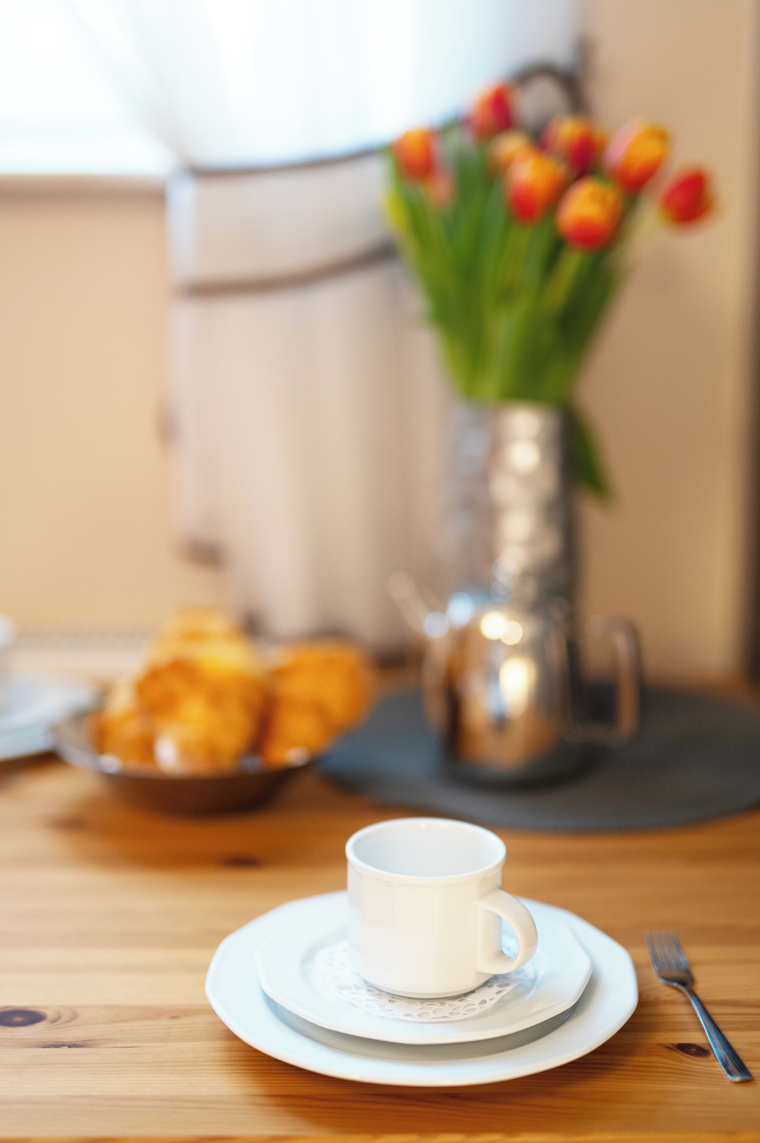 A table with a white cup and saucer, a fork, a bowl of pastries, and a vase with tulips.