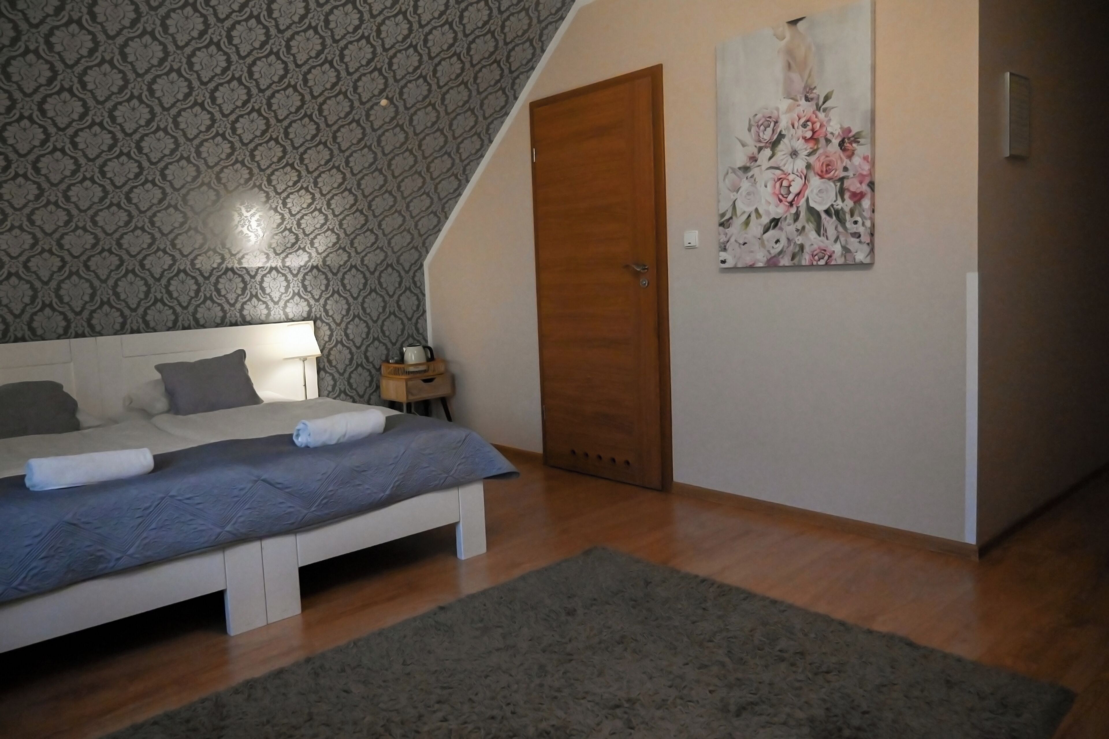 A bedroom with a bed, a nightstand, a wooden door, and a floral painting on the wall.